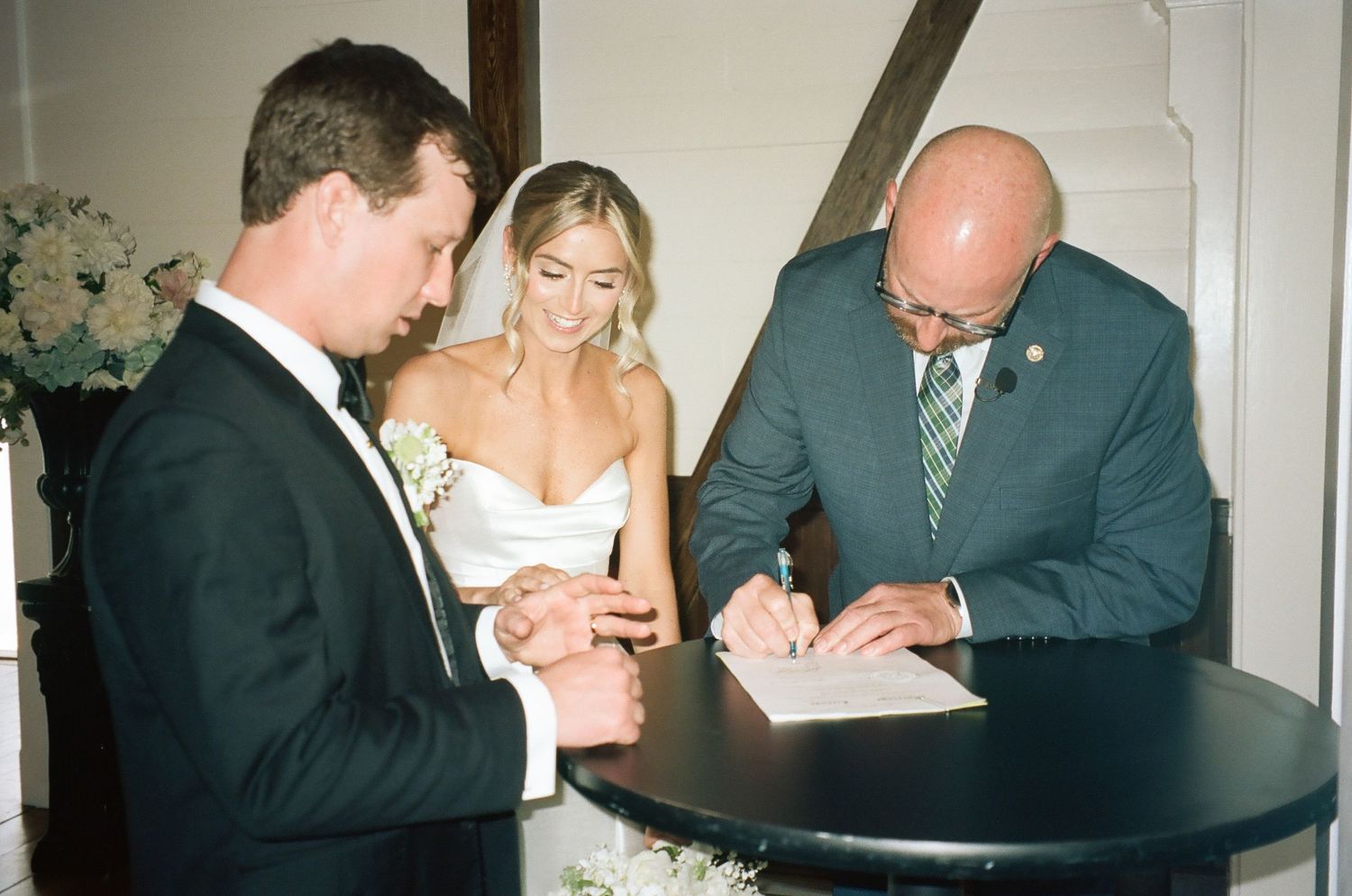 Bride and groom signing wedding certificate with officiant at elegant ceremony table with floral arrangements.