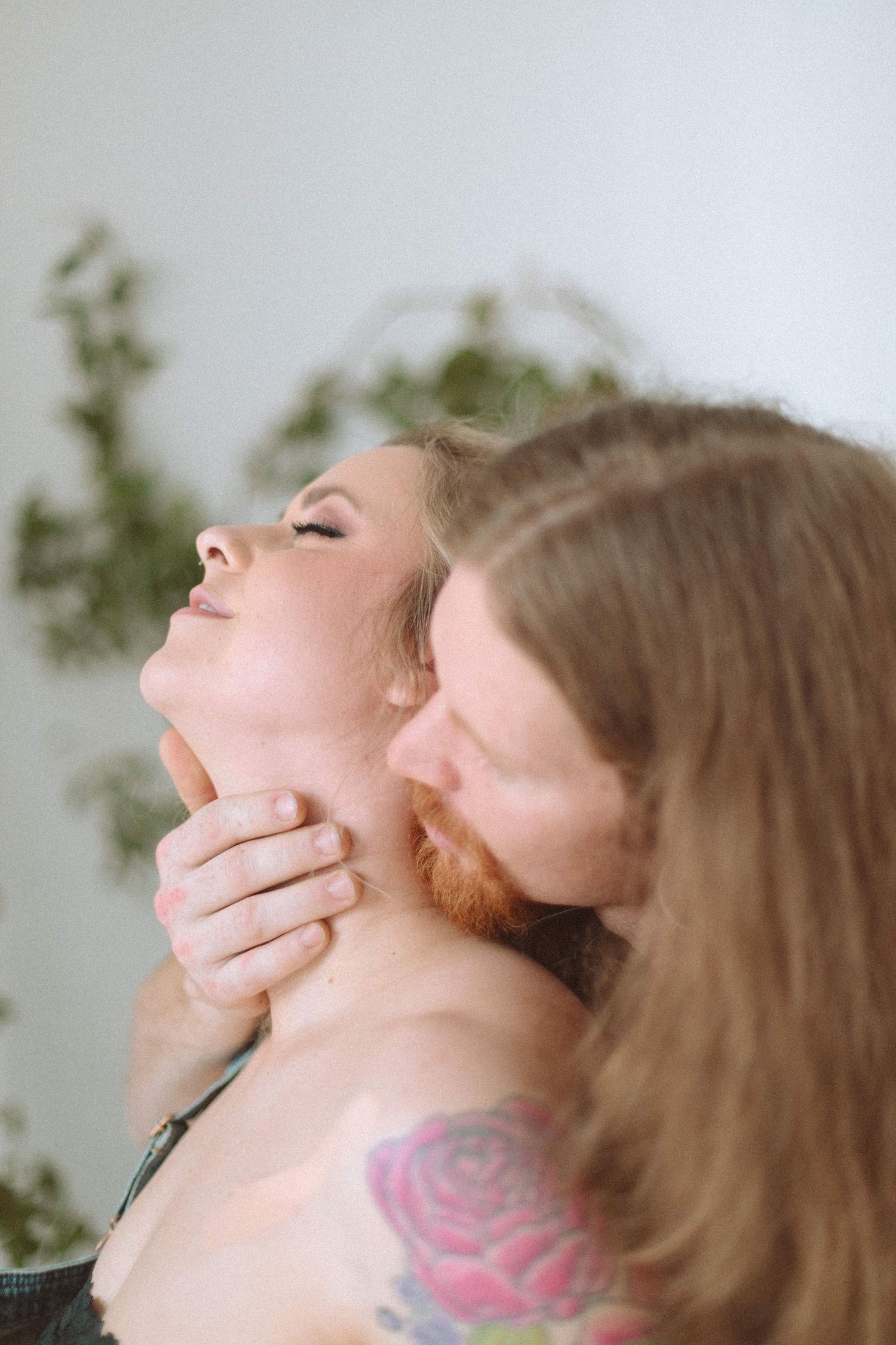 Intimate couple moment with tattooed woman tilting head back while partner embraces her from behind indoors.
