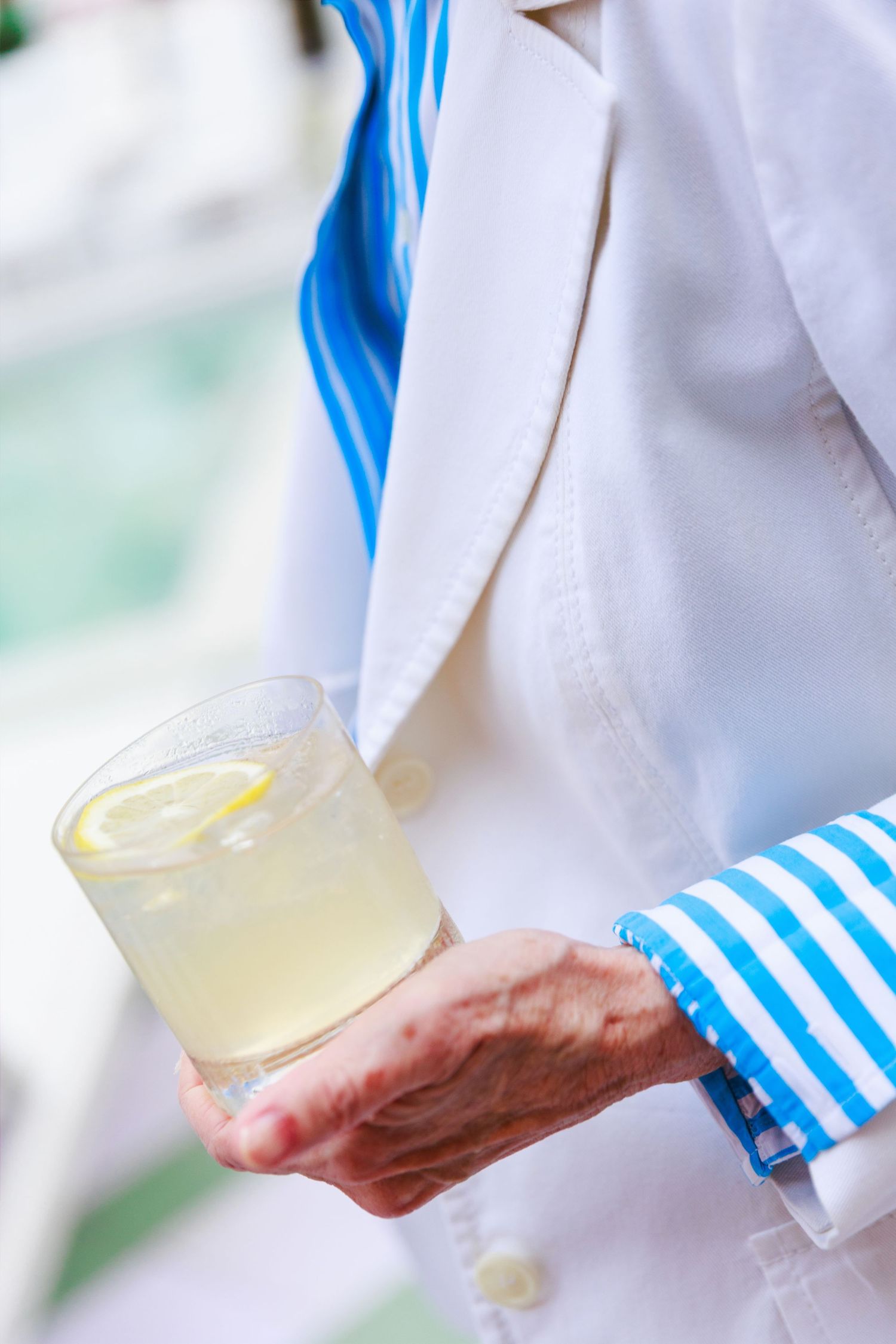 Elderly hand holding a glass of lemonade with ice and lemon slice, wearing blue striped cuff and white jacket.