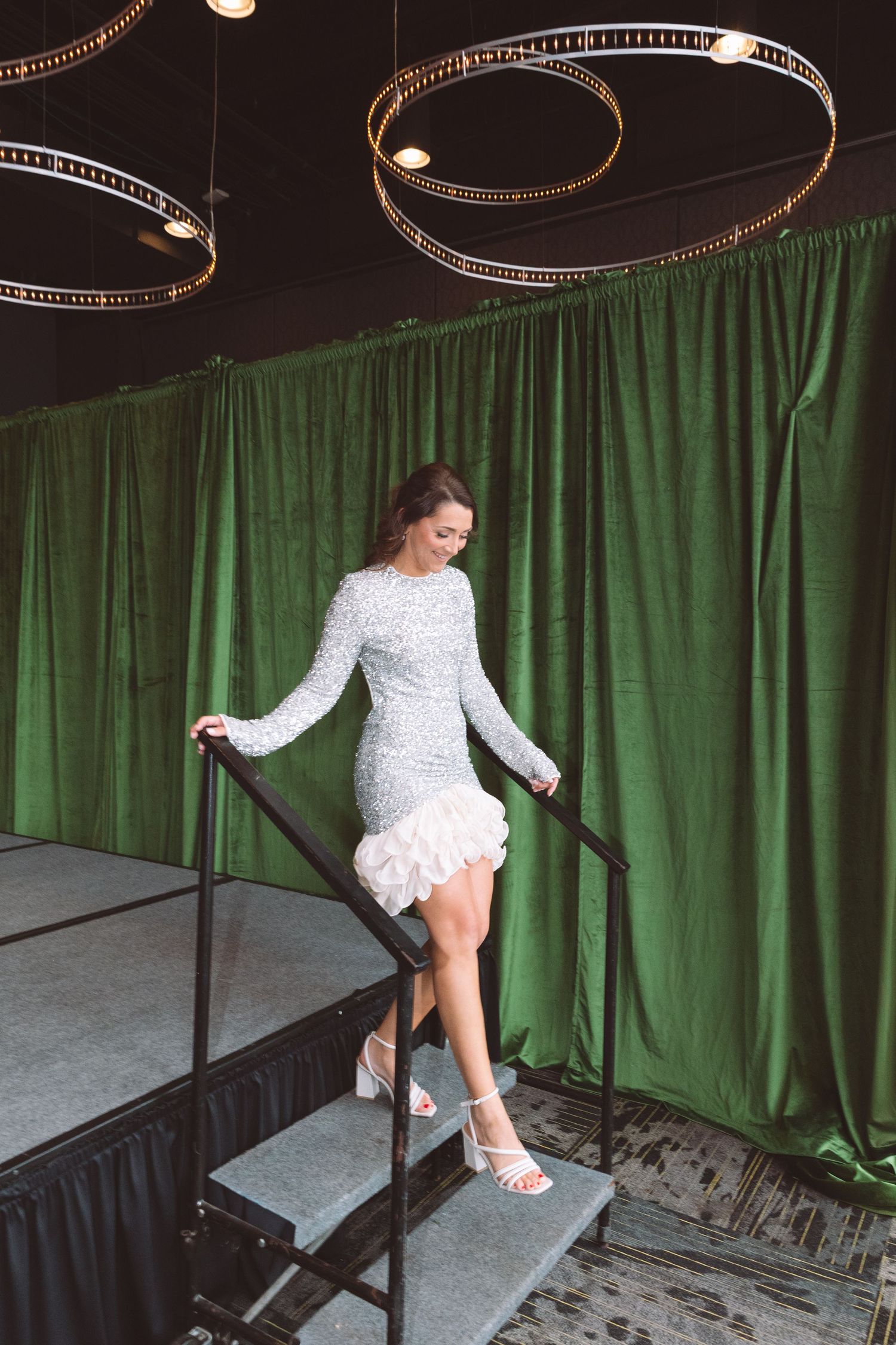 Woman in white sequin mini dress descending black stairs against green velvet curtain backdrop.