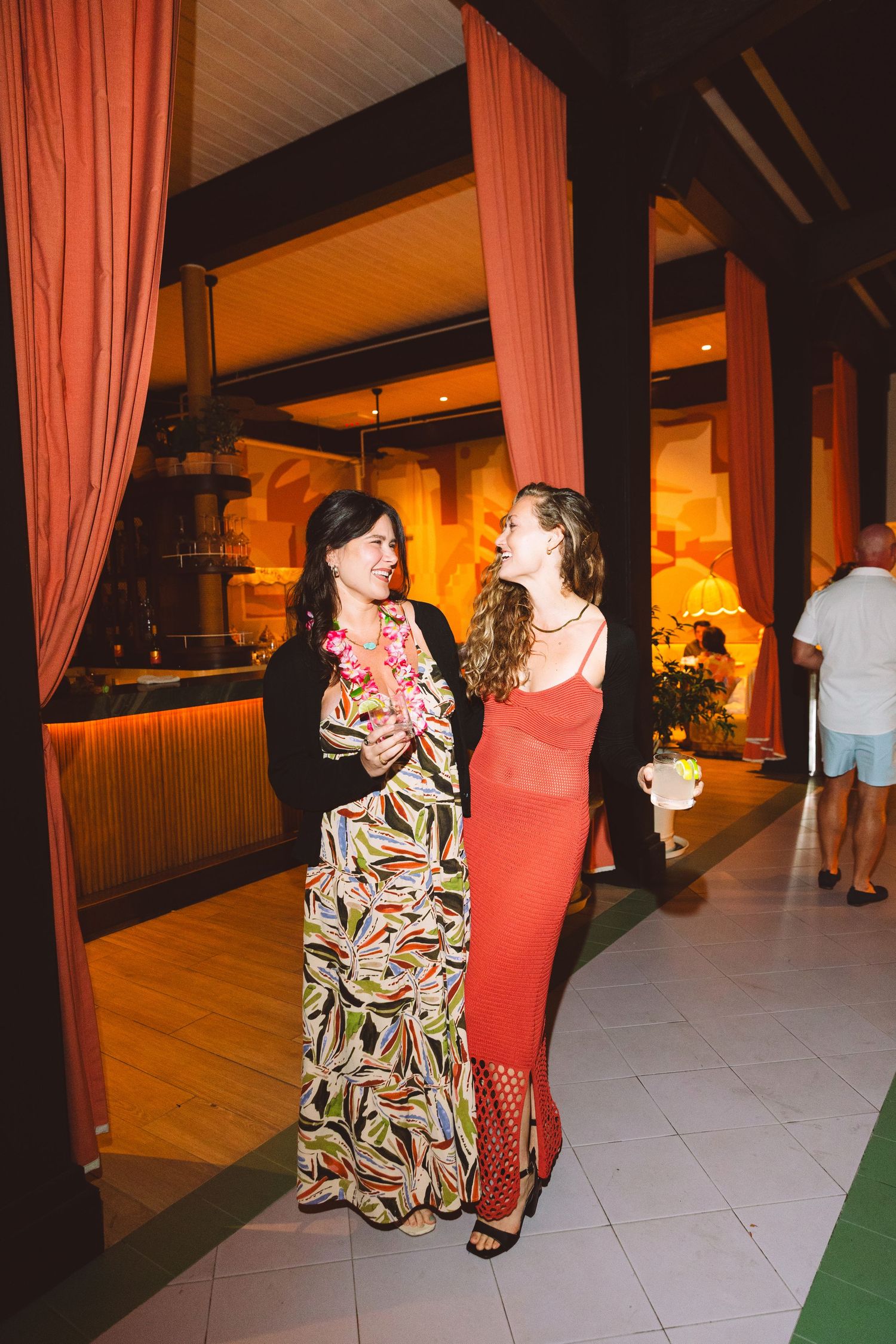 Two women in elegant dresses chat at an indoor party venue with pink curtains and warm lighting.