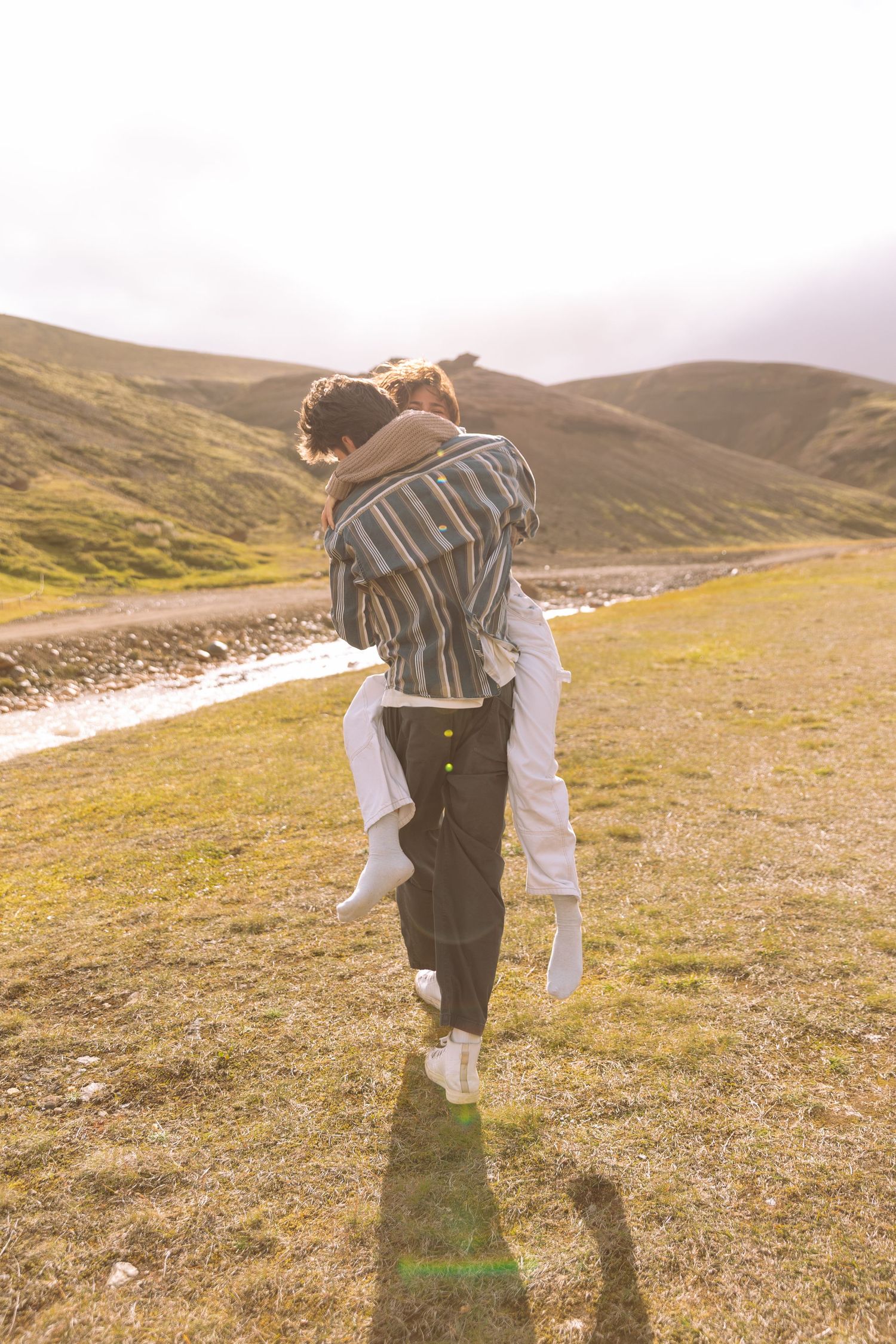 Man carrying woman on his back walking away toward a sunlit river valley in Iceland.