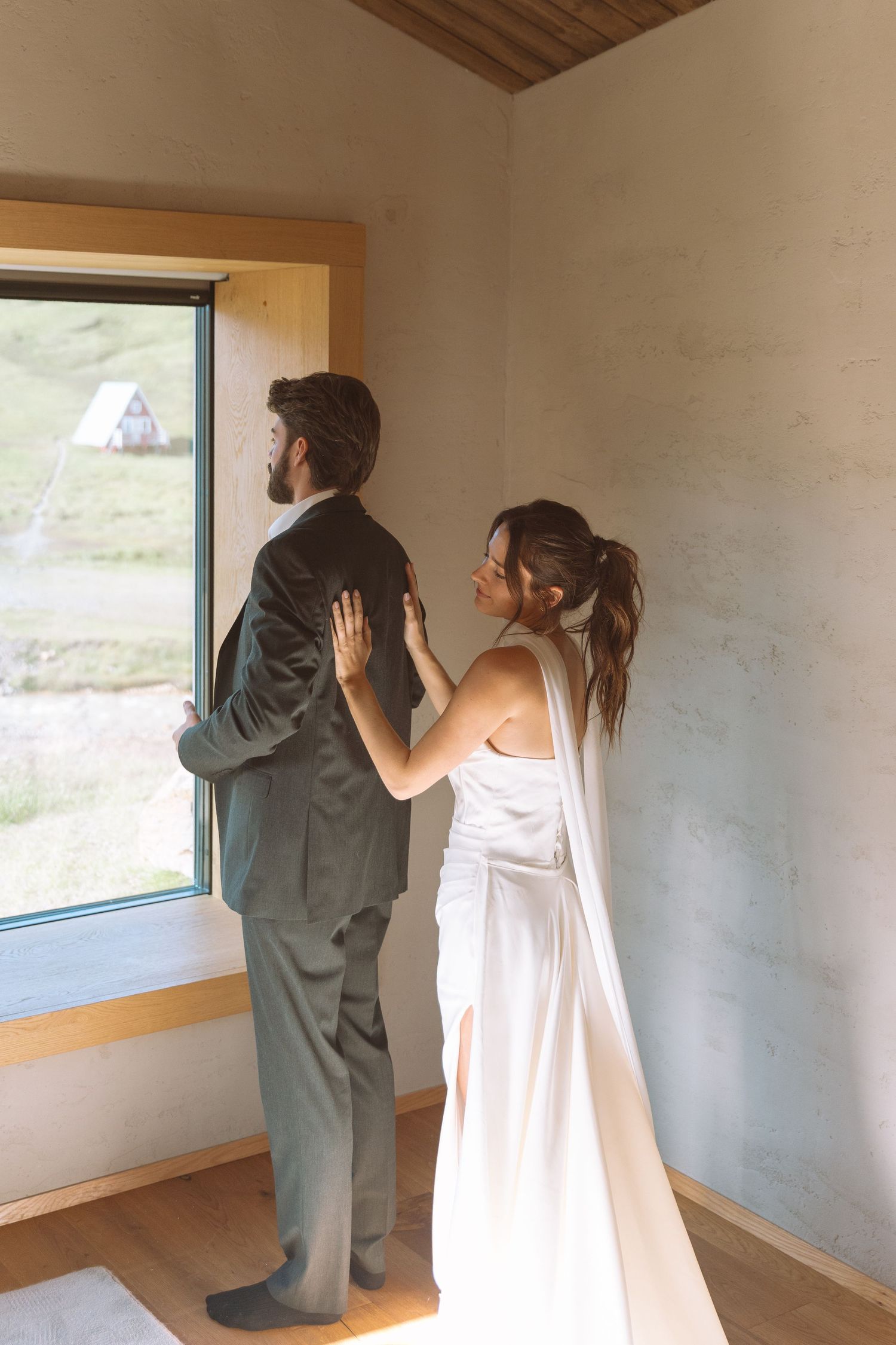 Bride gently placing hands on grooms back as they stand together by a window.
