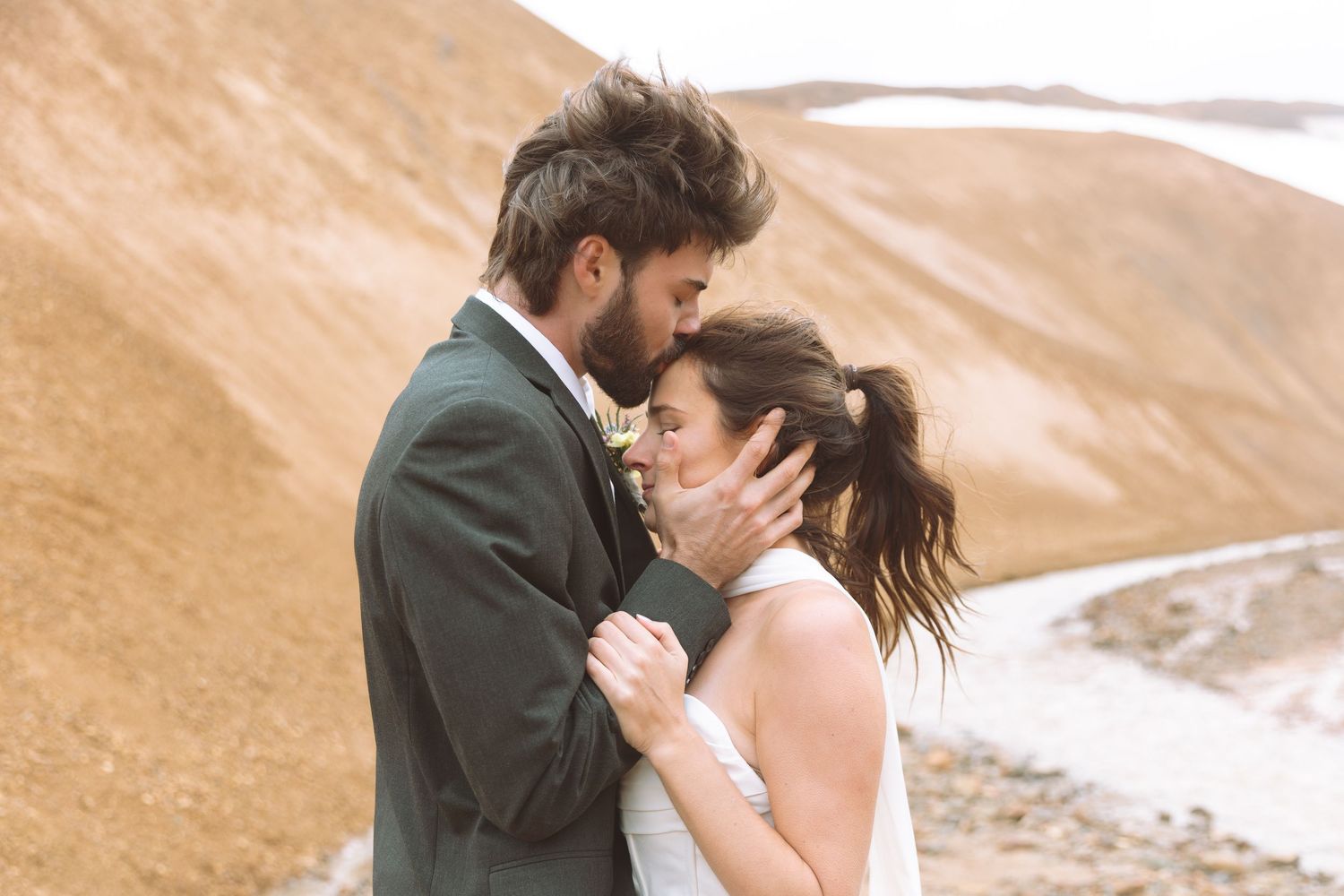 Bearded groom in dark suit tenderly embraces bride in white dress against rustic mountain backdrop.