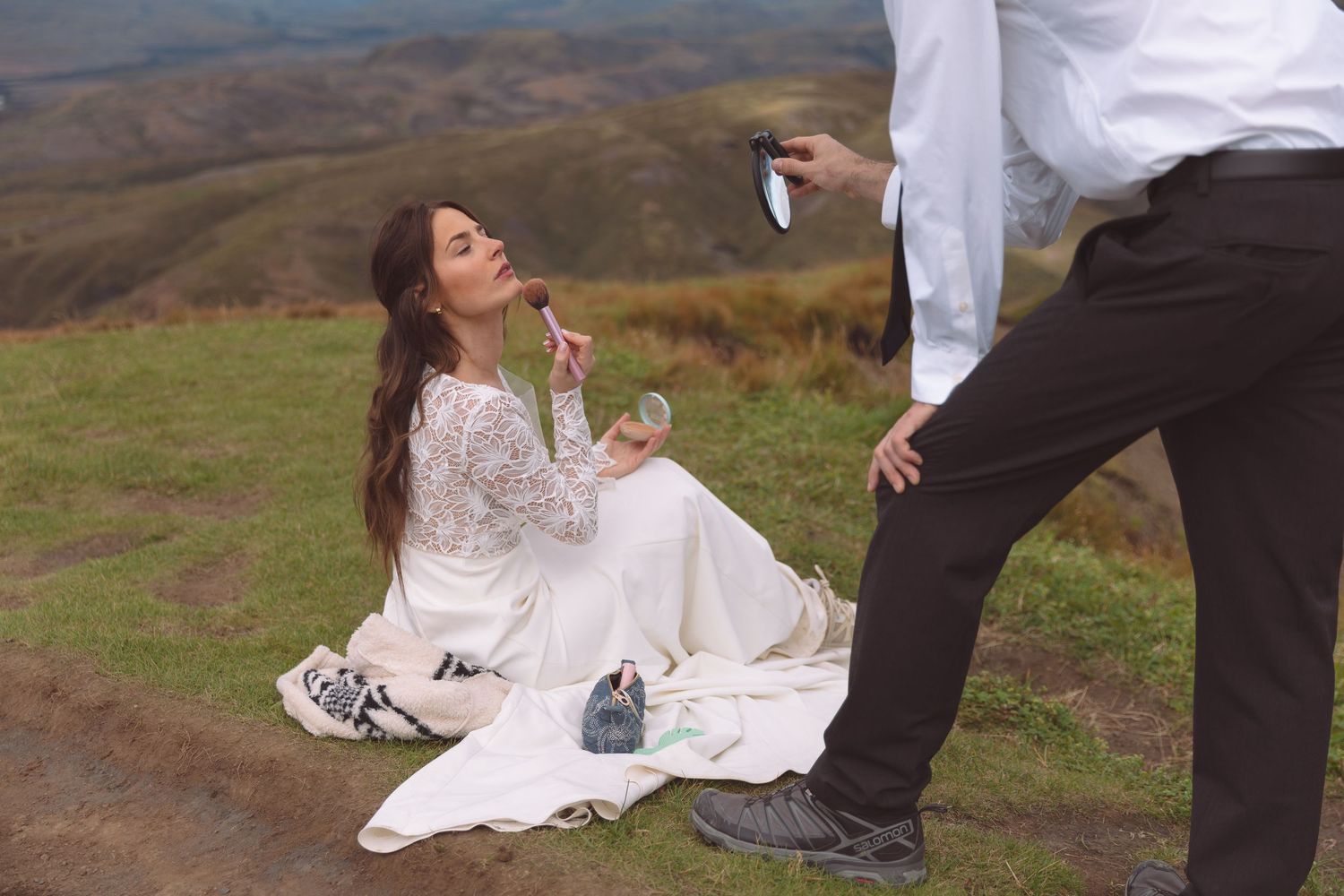 Bride touches up makeup with lipstick while groom holds mirror during scenic mountain wedding photoshoot.