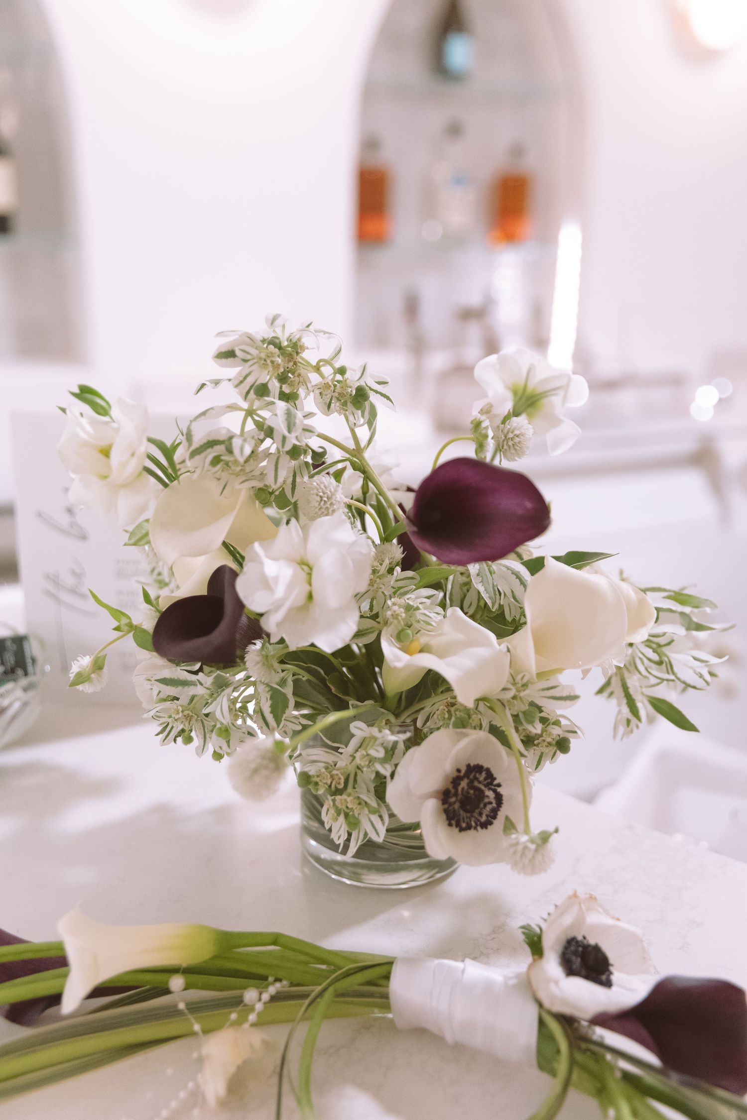 A elegant floral centerpiece featuring white anemones and deep purple calla lilies in a glass vase on a white table.