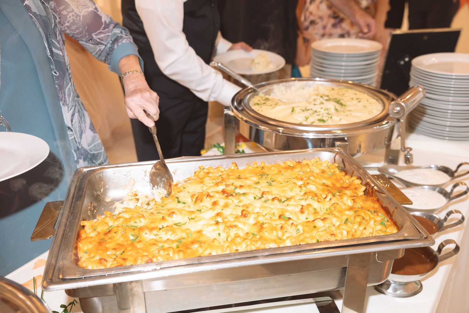 Guests serving cheesy casserole from a large rectangular chafing dish at a catered buffet.