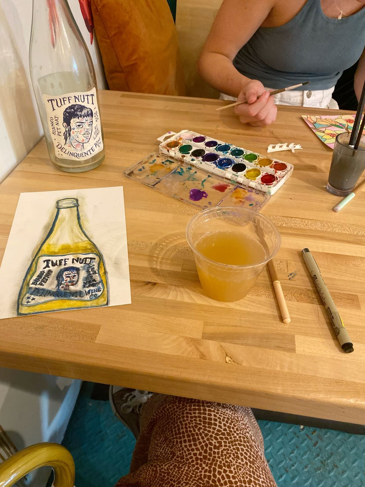 Watercolor painting of a Tuff Nutt bottle on a wooden table with paints, brushes, and a drink.