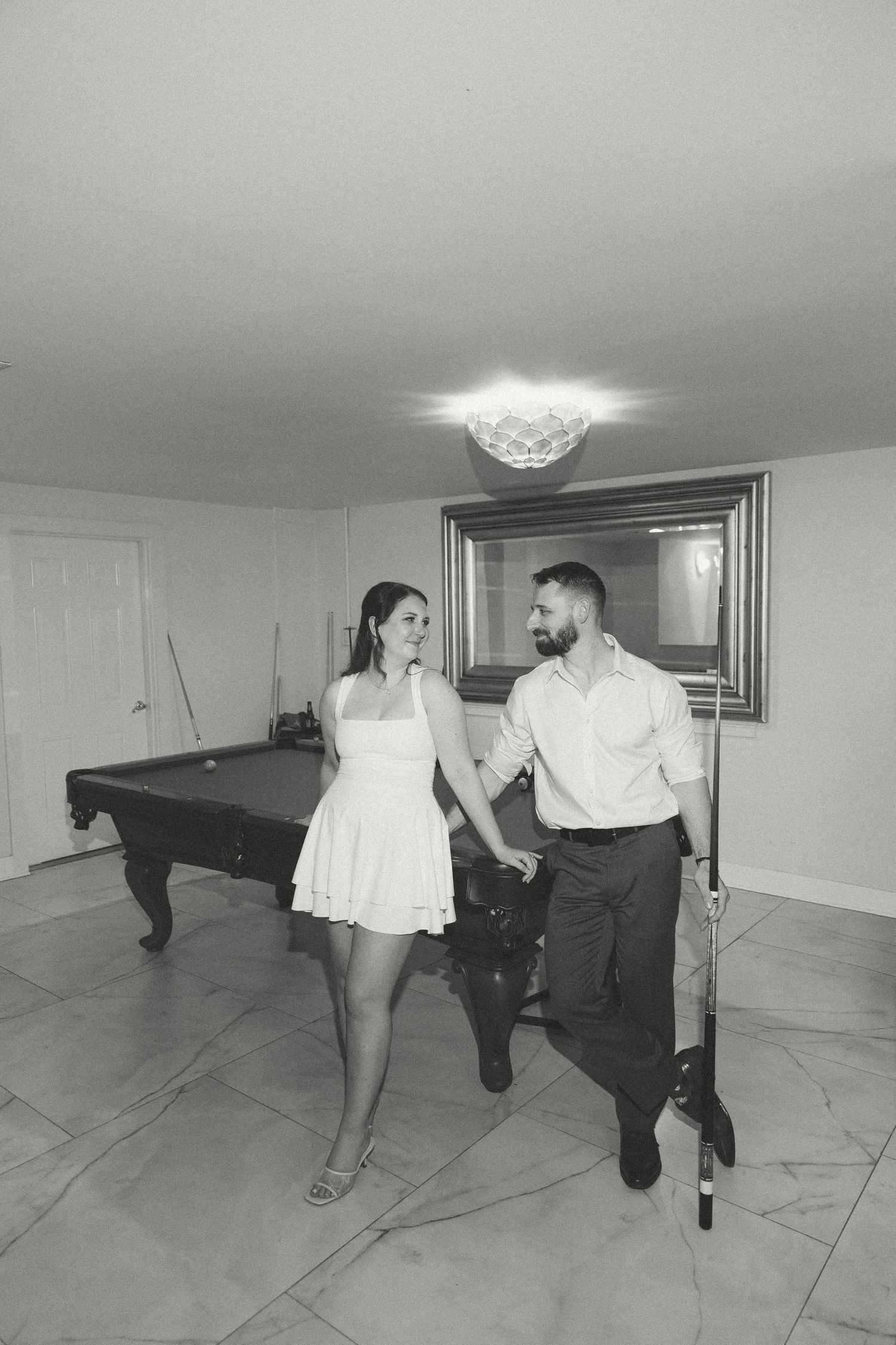 Black and white photo of a couple holding hands and smiling near a pool table in a game room.