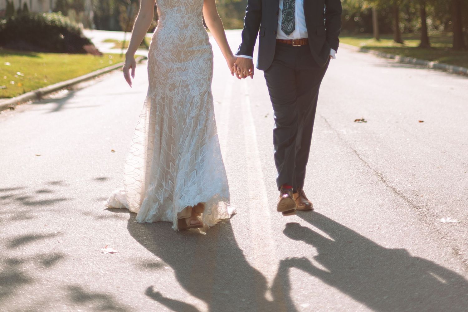 Bride and groom walk hand in hand down a sunlit road, casting long shadows on the pavement.
