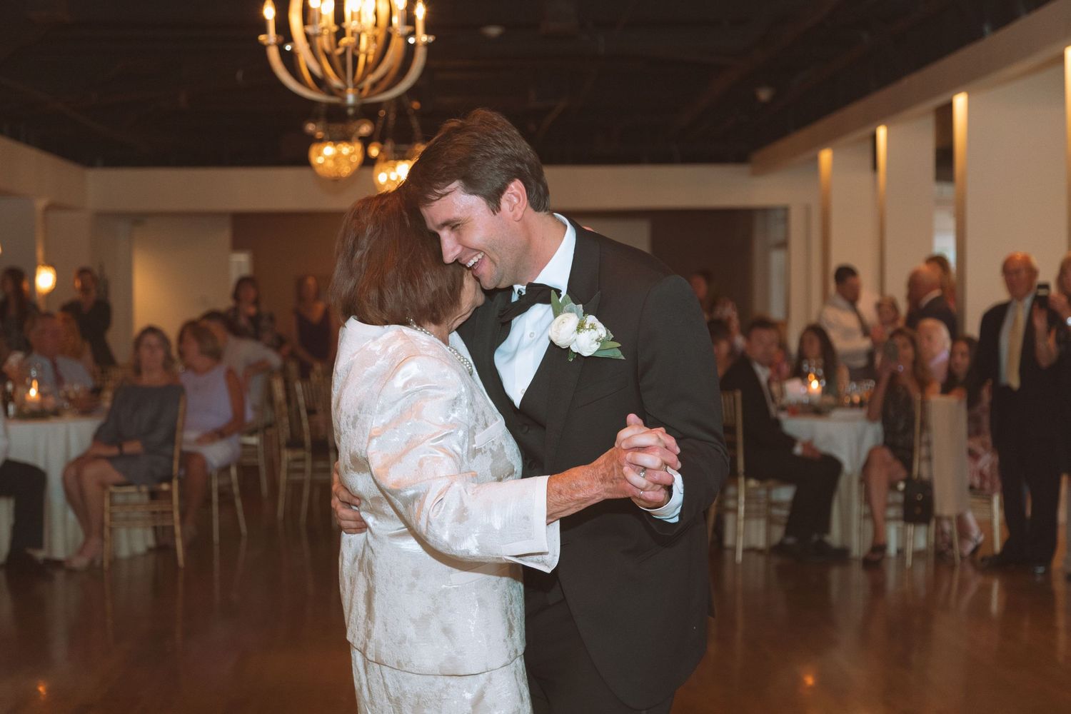 Groom sharing first dance with mother in elegant candlelit wedding reception venue.
