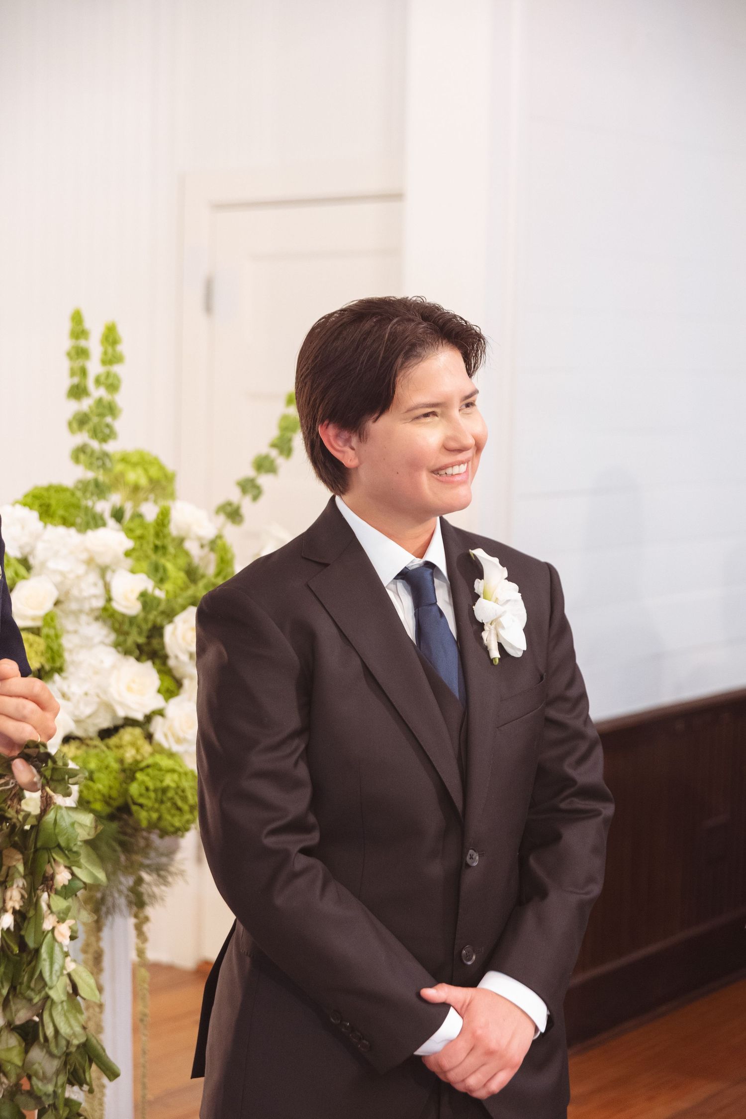 Smiling groom in dark suit with white boutonniere awaiting bride at wedding ceremony altar.