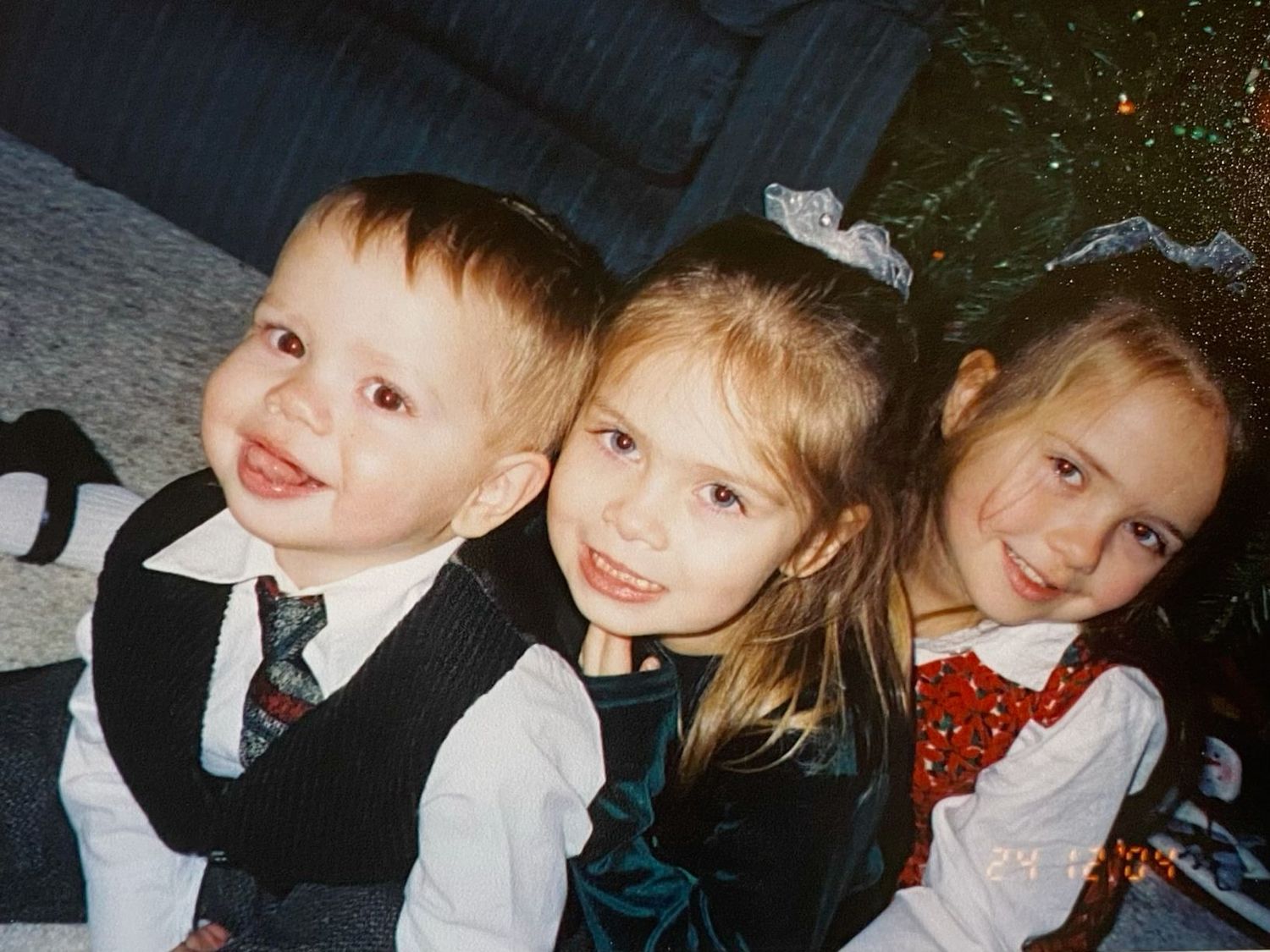 Three young children dressed in formal holiday attire smile together near a Christmas tree in a vintage photo.
