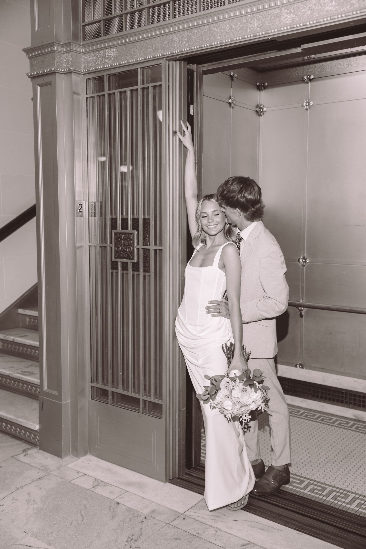 Black and white photo of a bride and groom sharing a kiss in an elegant elevator doorway, bride holding a bouquet.