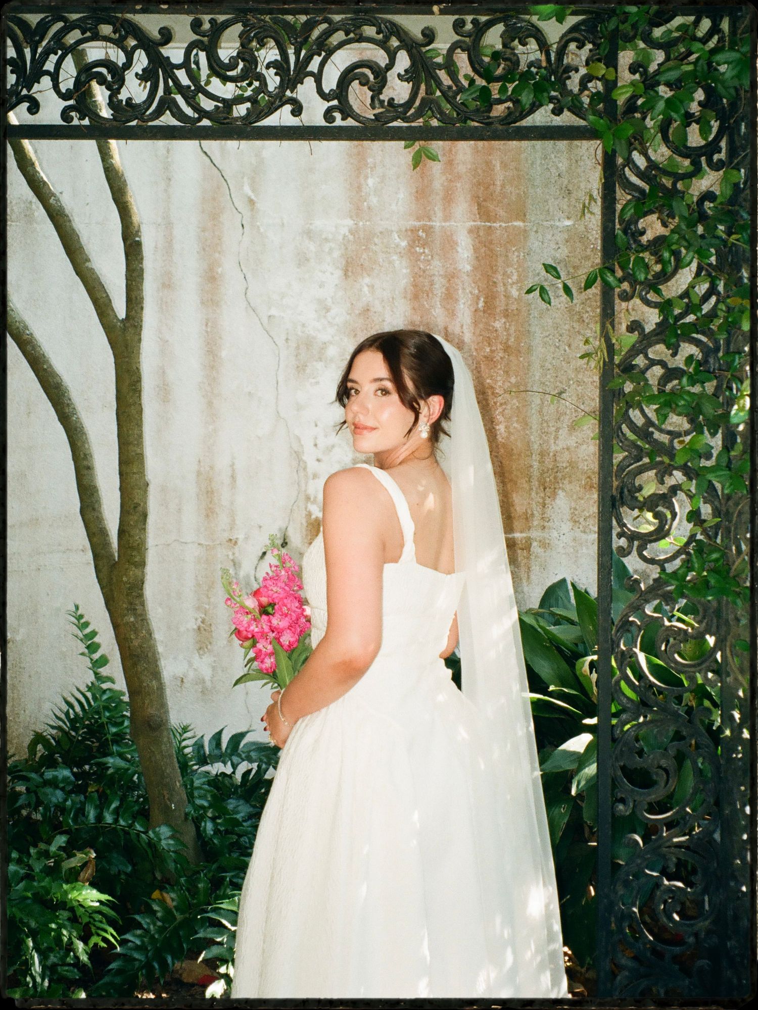Bride in a white gown and veil holding a pink bouquet framed by ornate black ironwork gate.