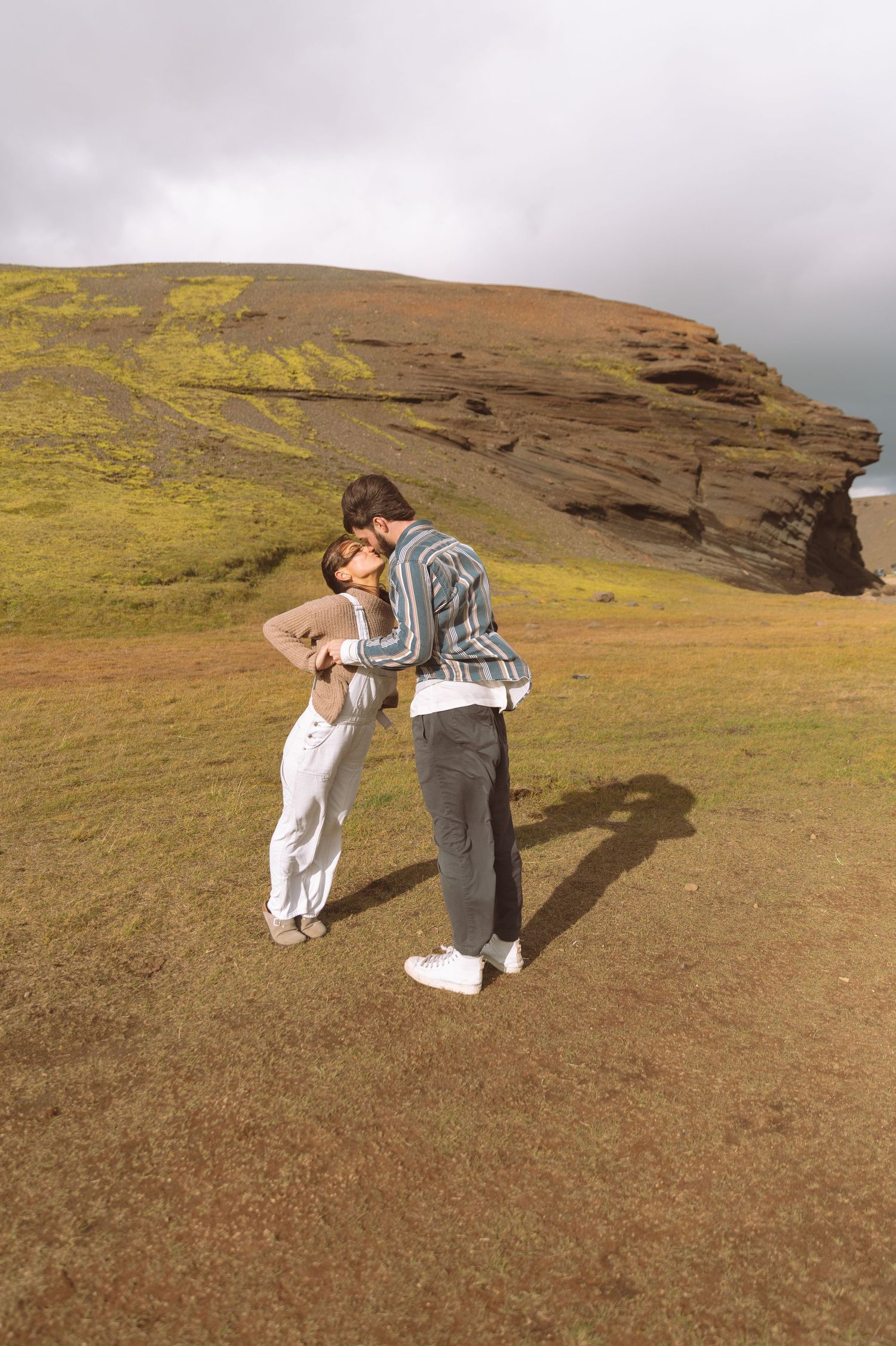 Romantic couple sharing a tender kiss on a grassy field with dramatic rocky hills in Iceland.