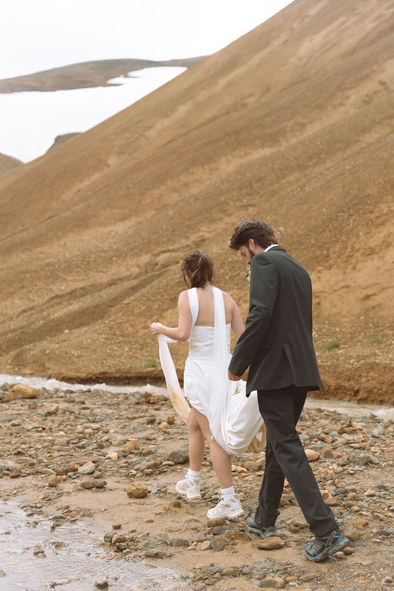 Bride lifts her white gown while groom assists her crossing a rocky stream during their mountain elopement.