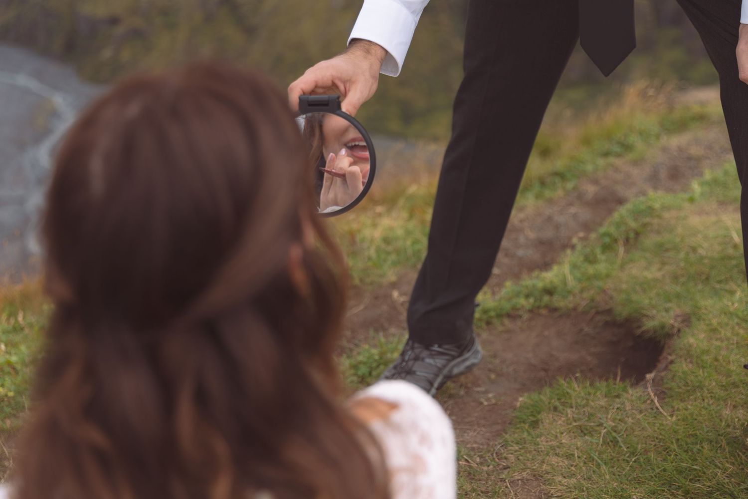 Groom holds small mirror reflecting bride's face during outdoor wedding photoshoot on grassy hillside.