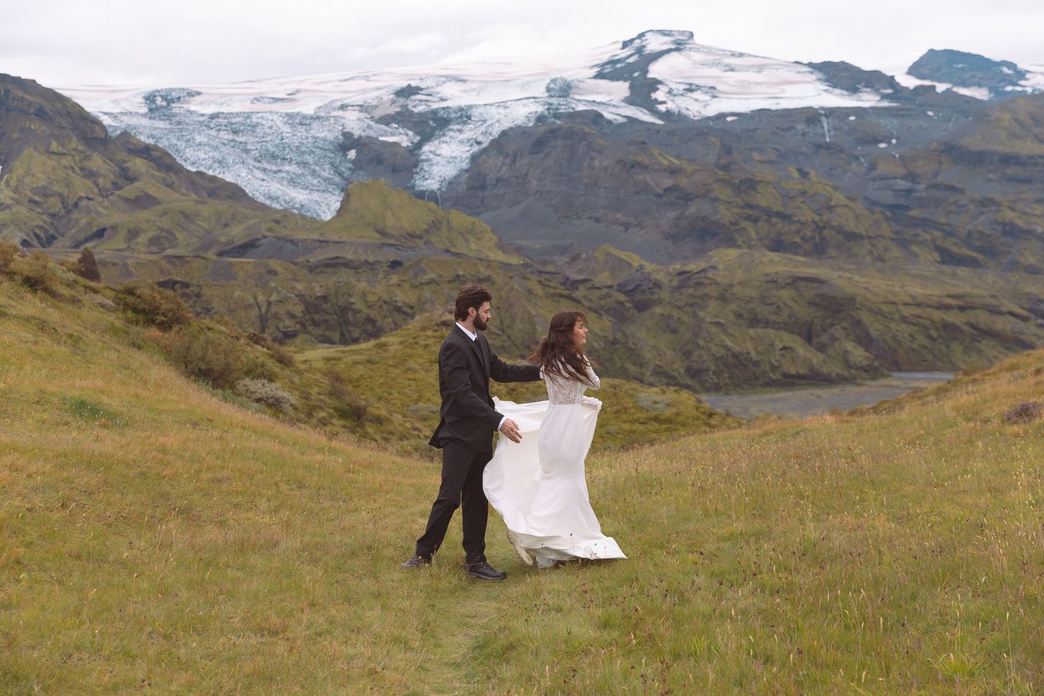 Newlyweds walking hand in hand across lush green Icelandic landscape with glacier views.