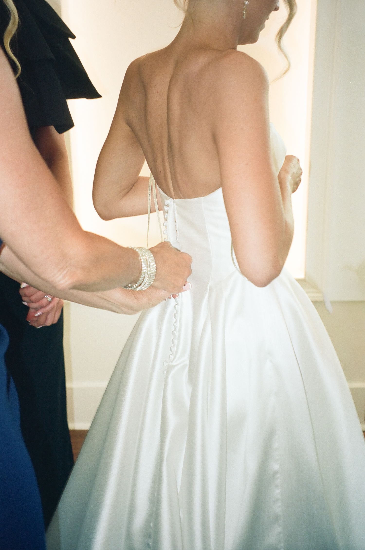 Helper lacing up the back of a strapless white satin wedding gown on a bride.