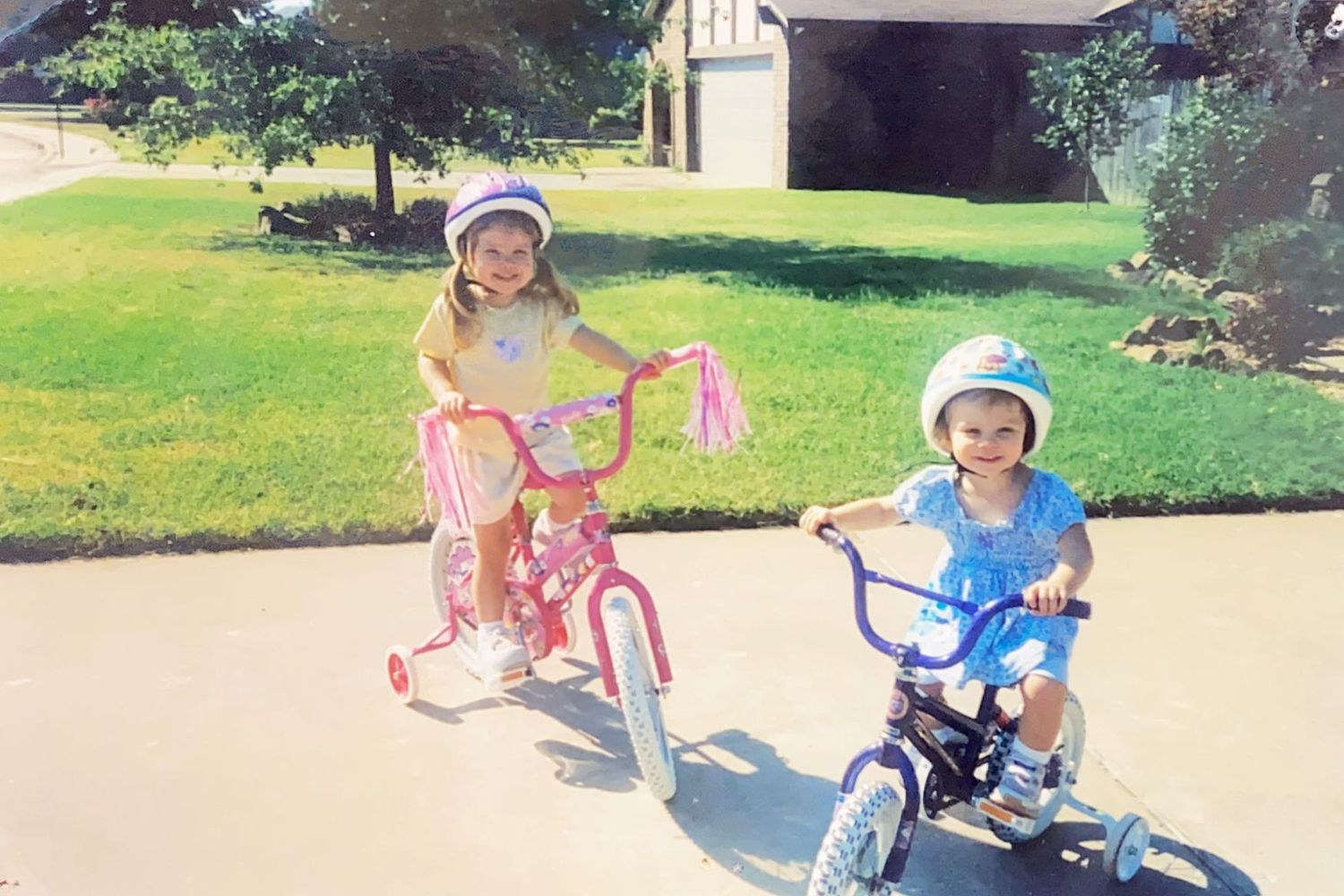 Two young girls ride bikes with training wheels and helmets on a sunny driveway.