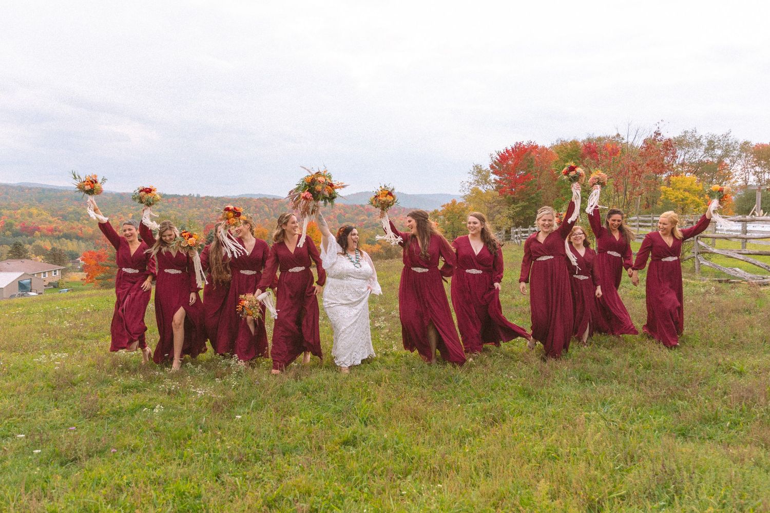 Bridesmaids in burgundy dresses joyfully raise bouquets of autumn flowers on a hillside with vibrant fall foliage backdrop.