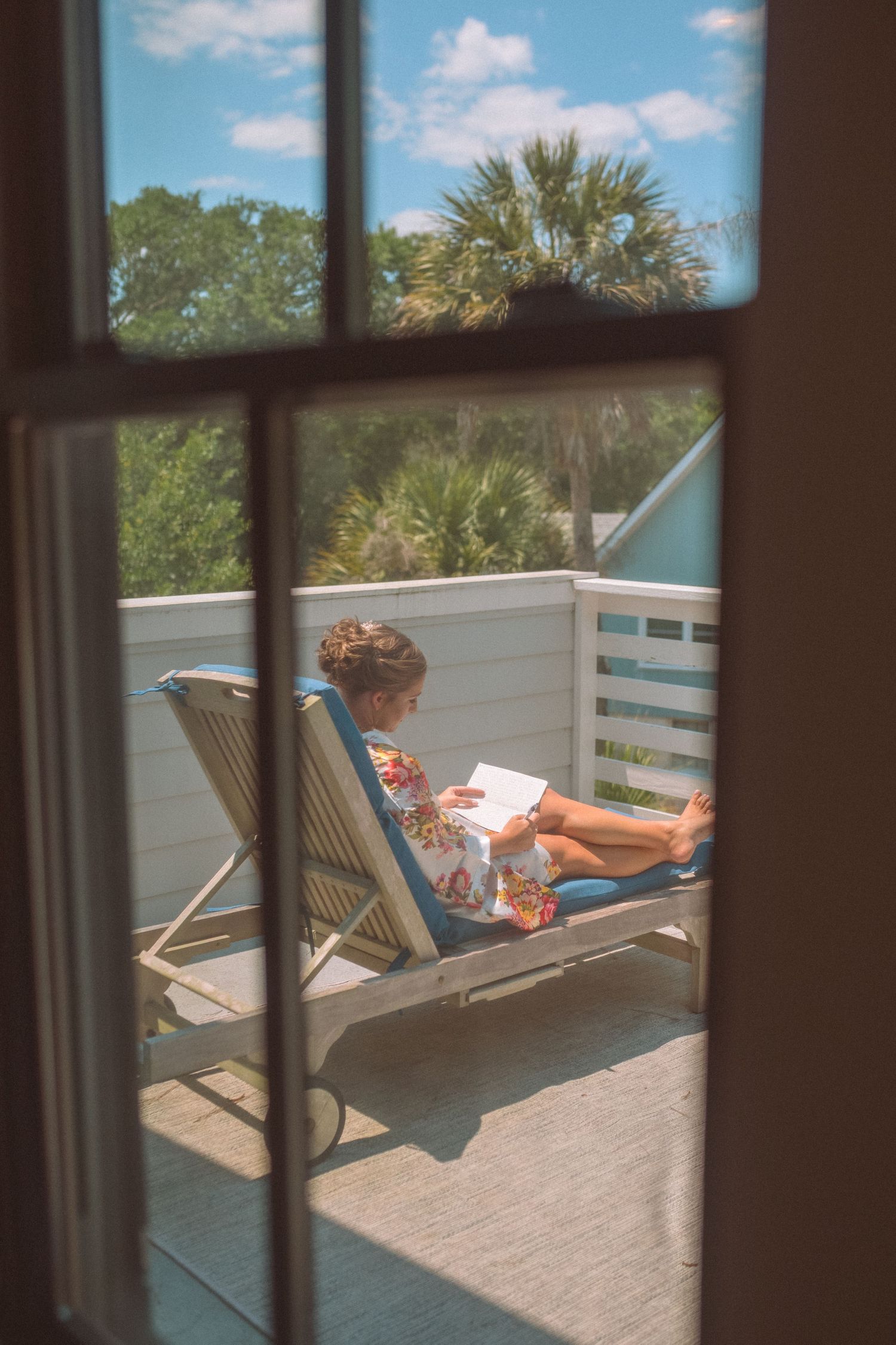 Bride in floral robe relaxing on lounge chair reading on sunny outdoor deck with palm trees.