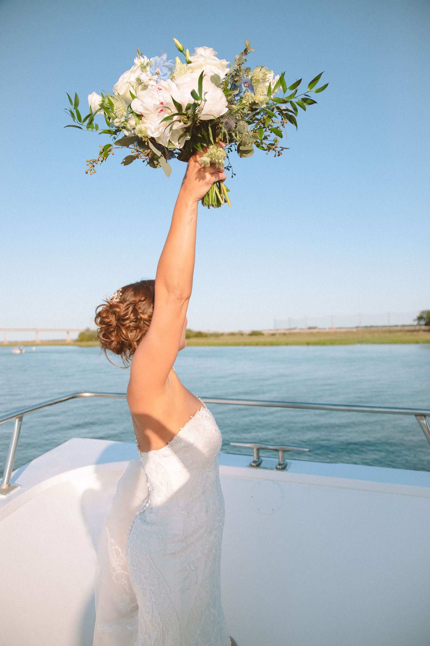 A bride in a white lace gown triumphantly raises her bouquet of white flowers on a yacht deck.