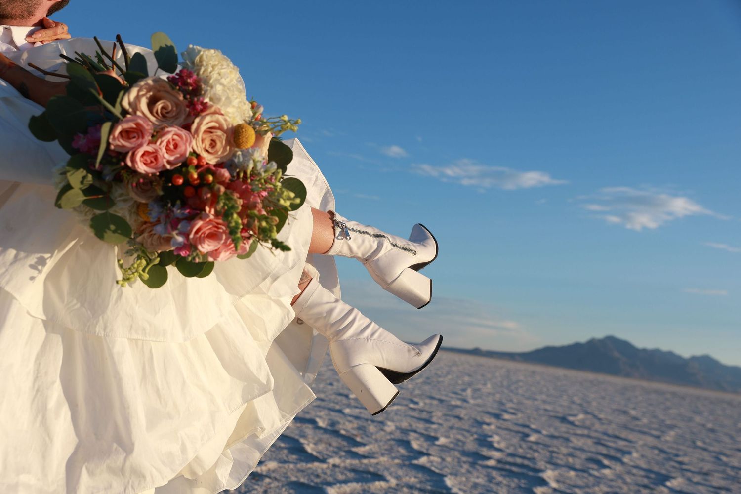 Bride in white dress and white boots holding a colorful bouquet at Bonneville Salt Flats with mountains.