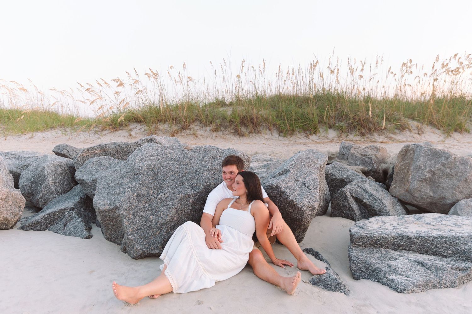 A couple in white outfits sits romantically against large rocks on a sandy beach with sea oats in the background.