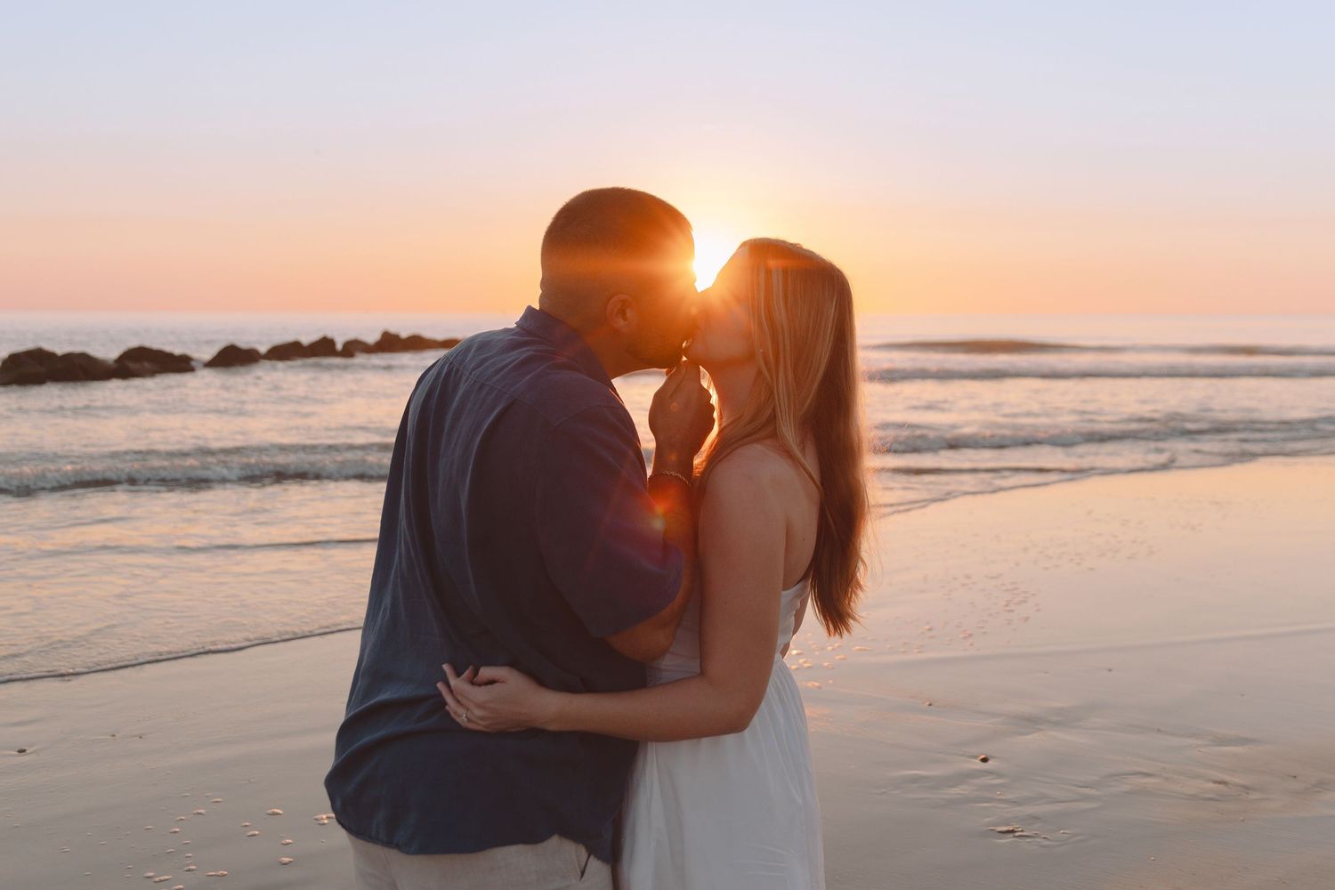 Couple sharing a romantic kiss on the beach at golden sunset with ocean waves in the background.