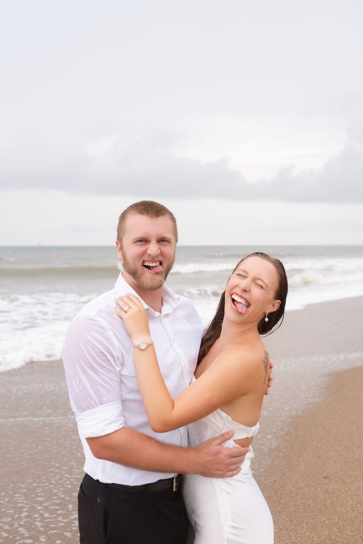 Joyful couple laughing and embracing on a beach, both making silly faces with ocean waves behind them.