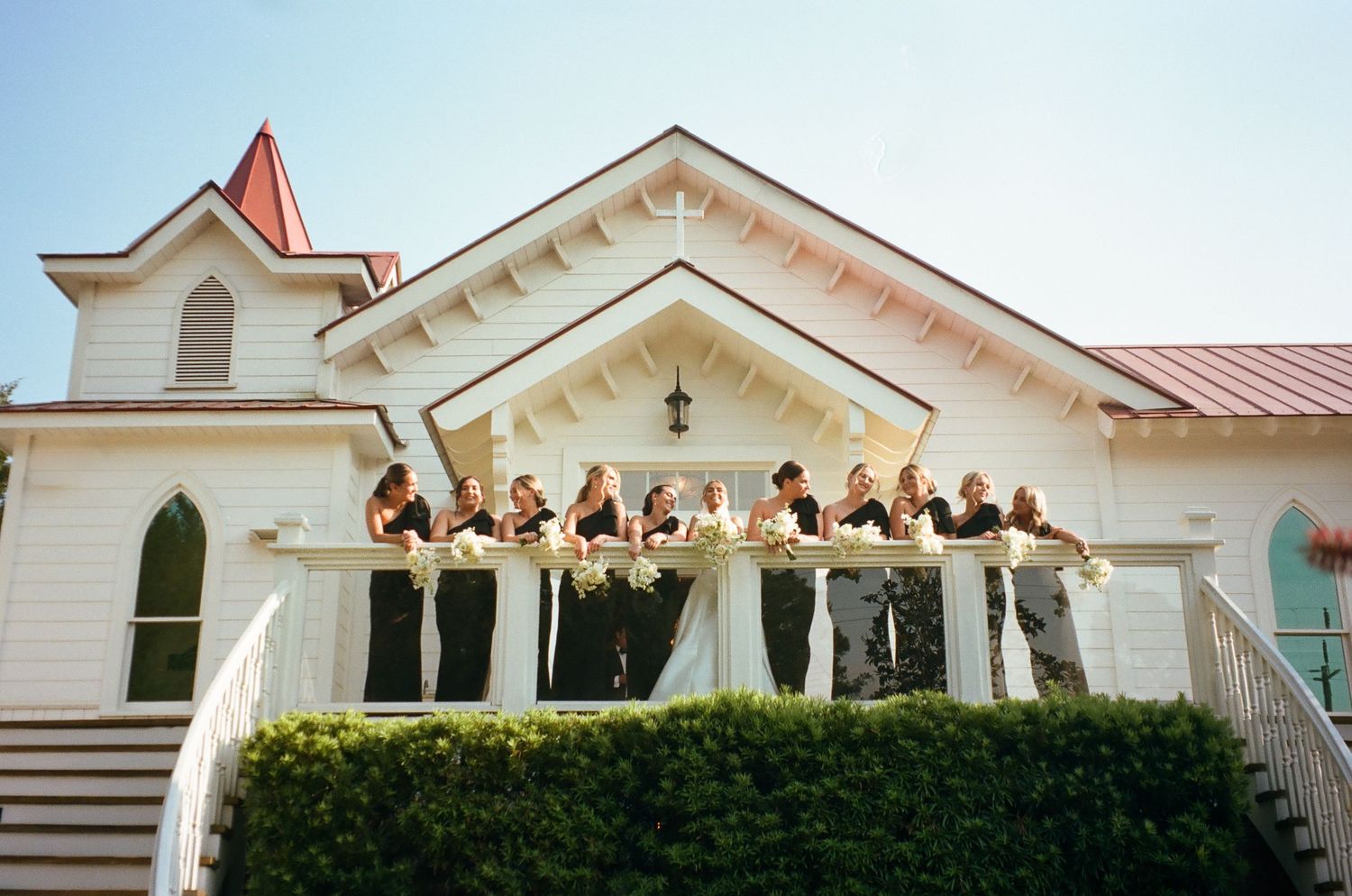 Wedding party poses on white chapel balcony adorned with floral garlands on a sunny day.