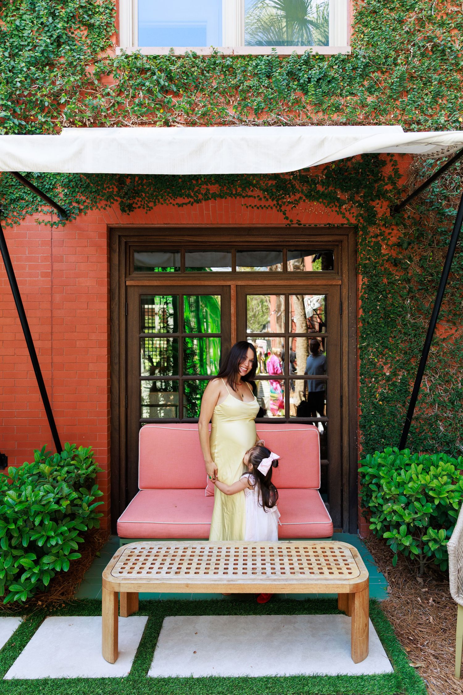 A woman in a yellow dress stands with a young girl on a pink outdoor sofa beside a ivy-covered brick building.