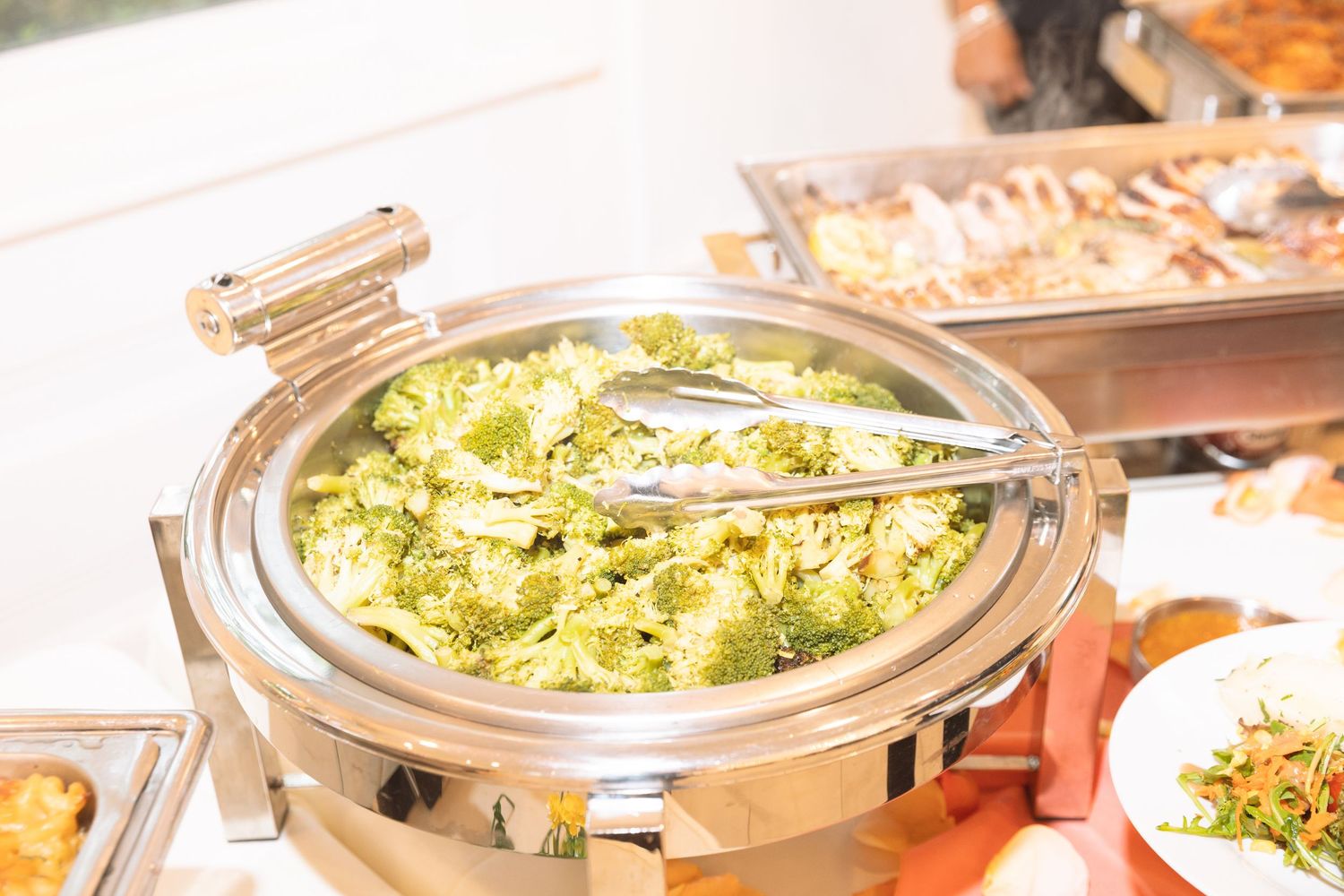 Round chafing dish filled with fresh broccoli and tongs at a catered buffet event.
