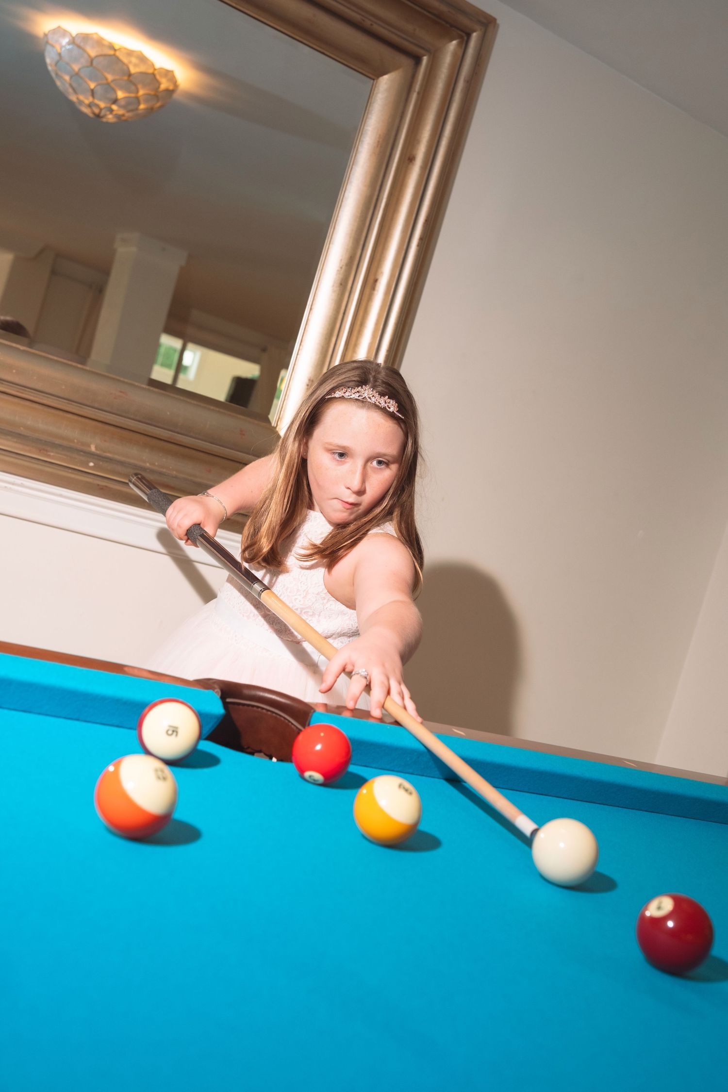 Young girl in white dress playing pool, leaning over blue billiard table with cue stick.