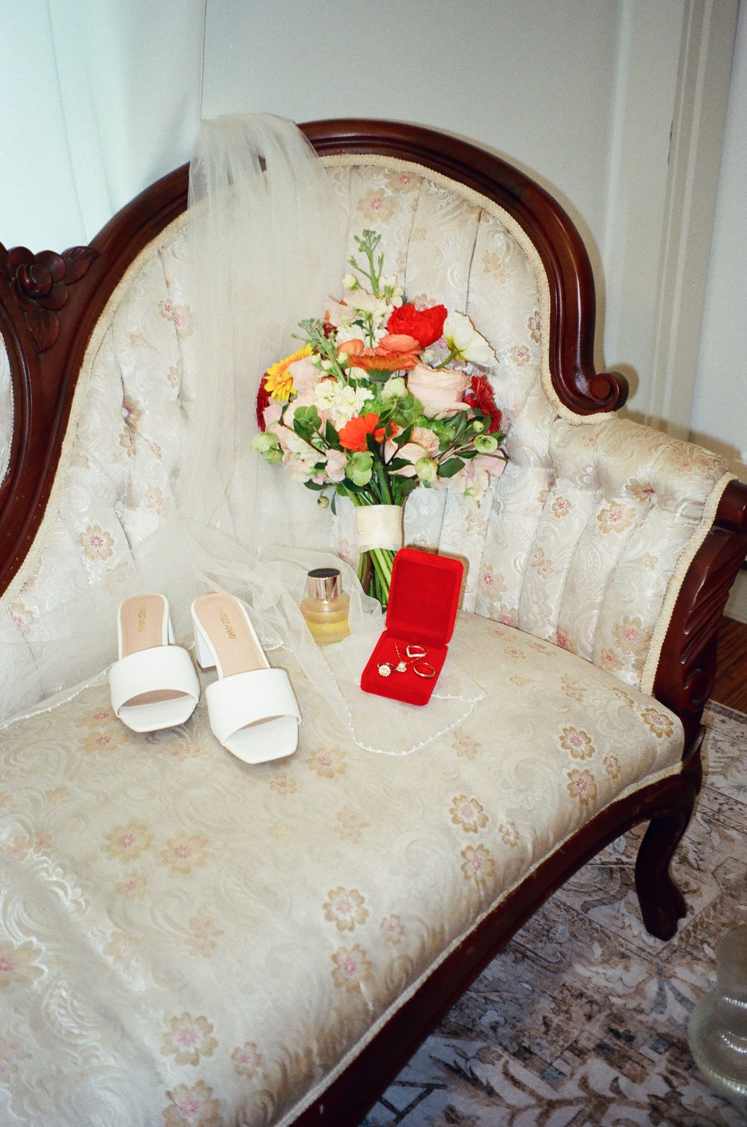 Elegant vintage sofa with bridal accessories including white heels, bouquet, perfume, and red jewelry box.
