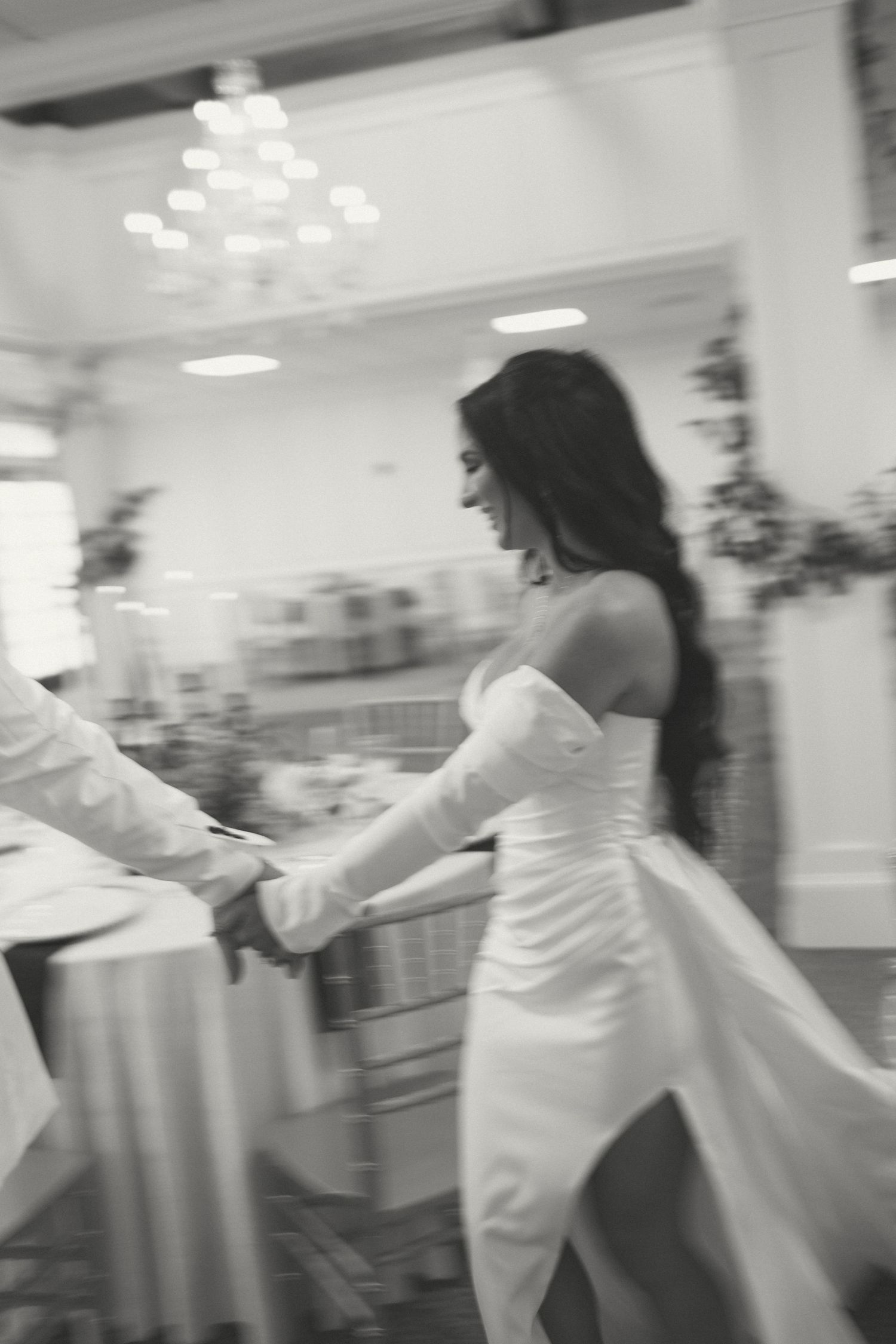 Black and white motion-blur photo of a smiling bride in a white gown walking through a chandelier-lit reception hall.