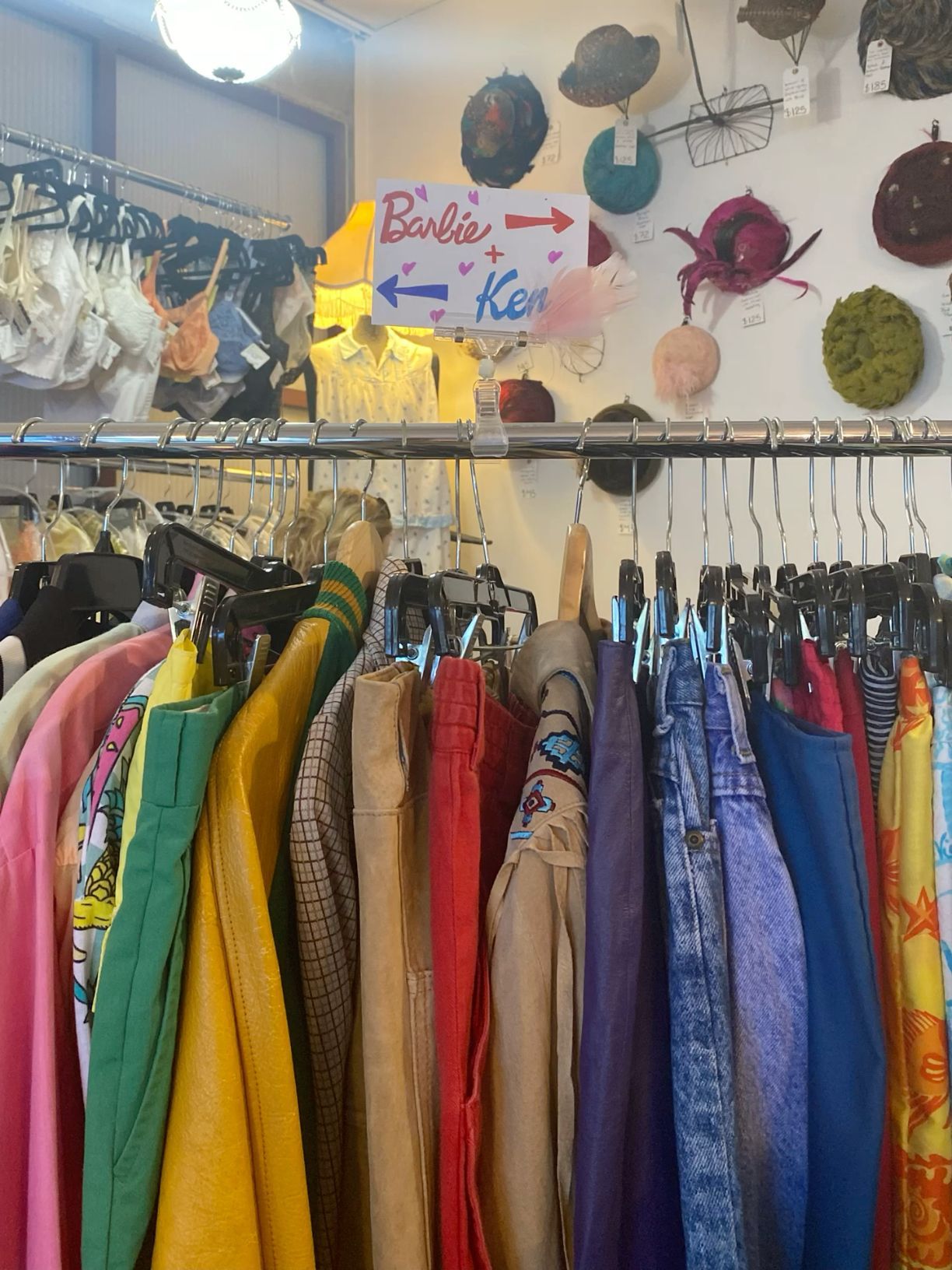 Colorful vintage clothing rack with Barbie and Ken sign and decorative hats displayed on wall behind it.