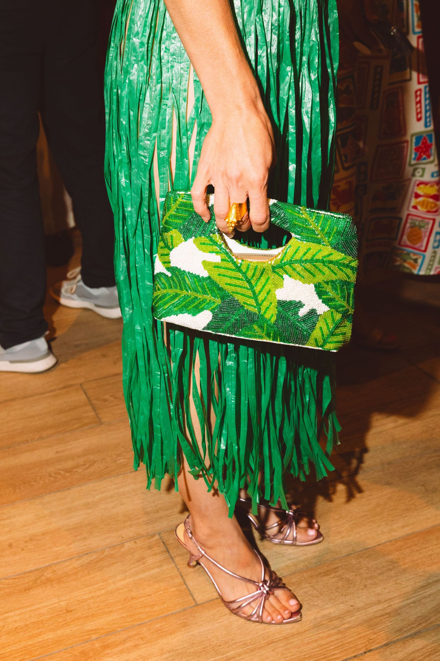 Woman holding green tropical leaf print clutch bag with fringe green sequin skirt and heels.