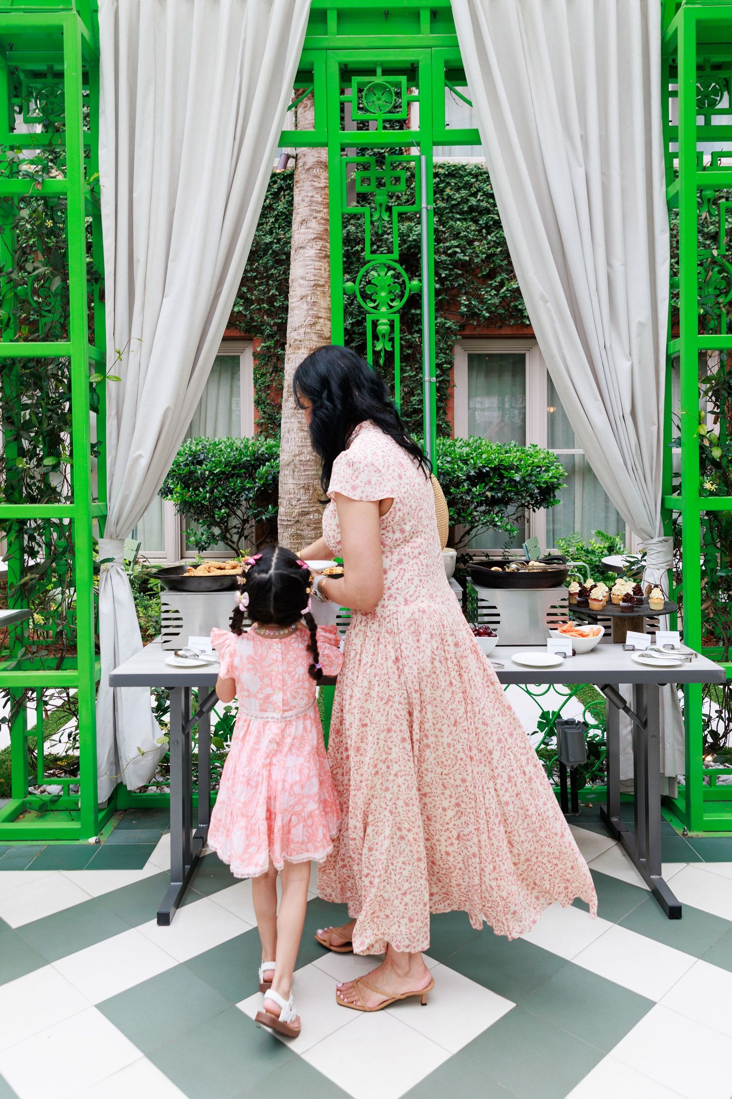 Mother and daughter in matching pink floral dresses stand at an outdoor dining table with green decorative panels.
