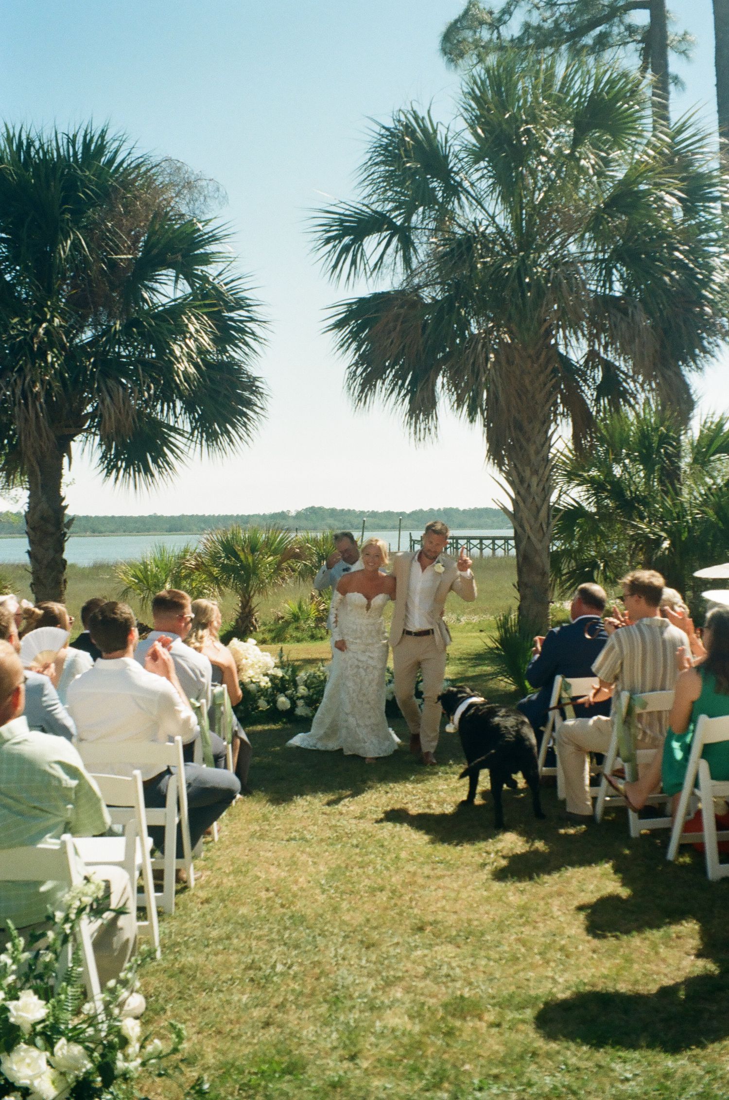 Outdoor waterfront wedding ceremony with bride and groom at altar, palm trees, and guests seated in white chairs.