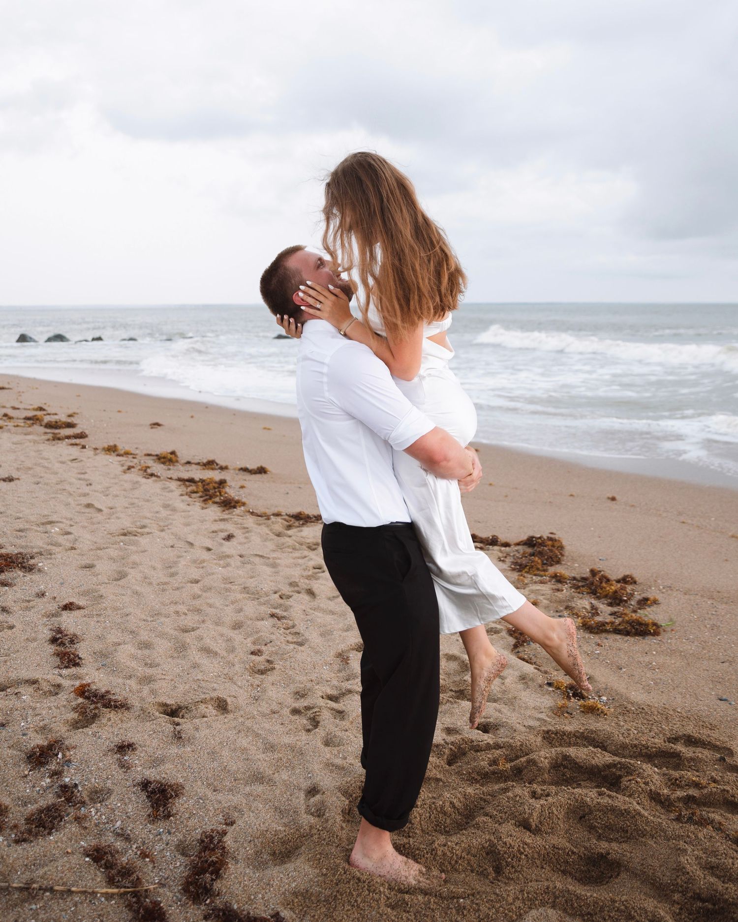 A man lifts a woman in a white dress for a kiss on a sandy beach.