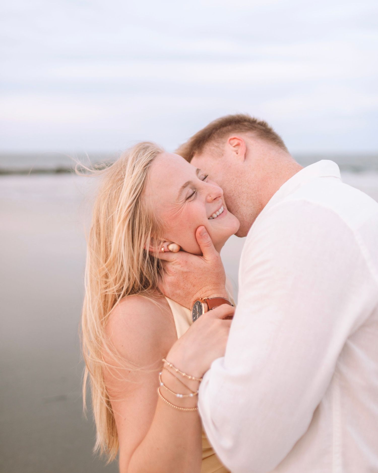 Man tenderly cupping a smiling blonde woman's face on a serene beach at dusk.