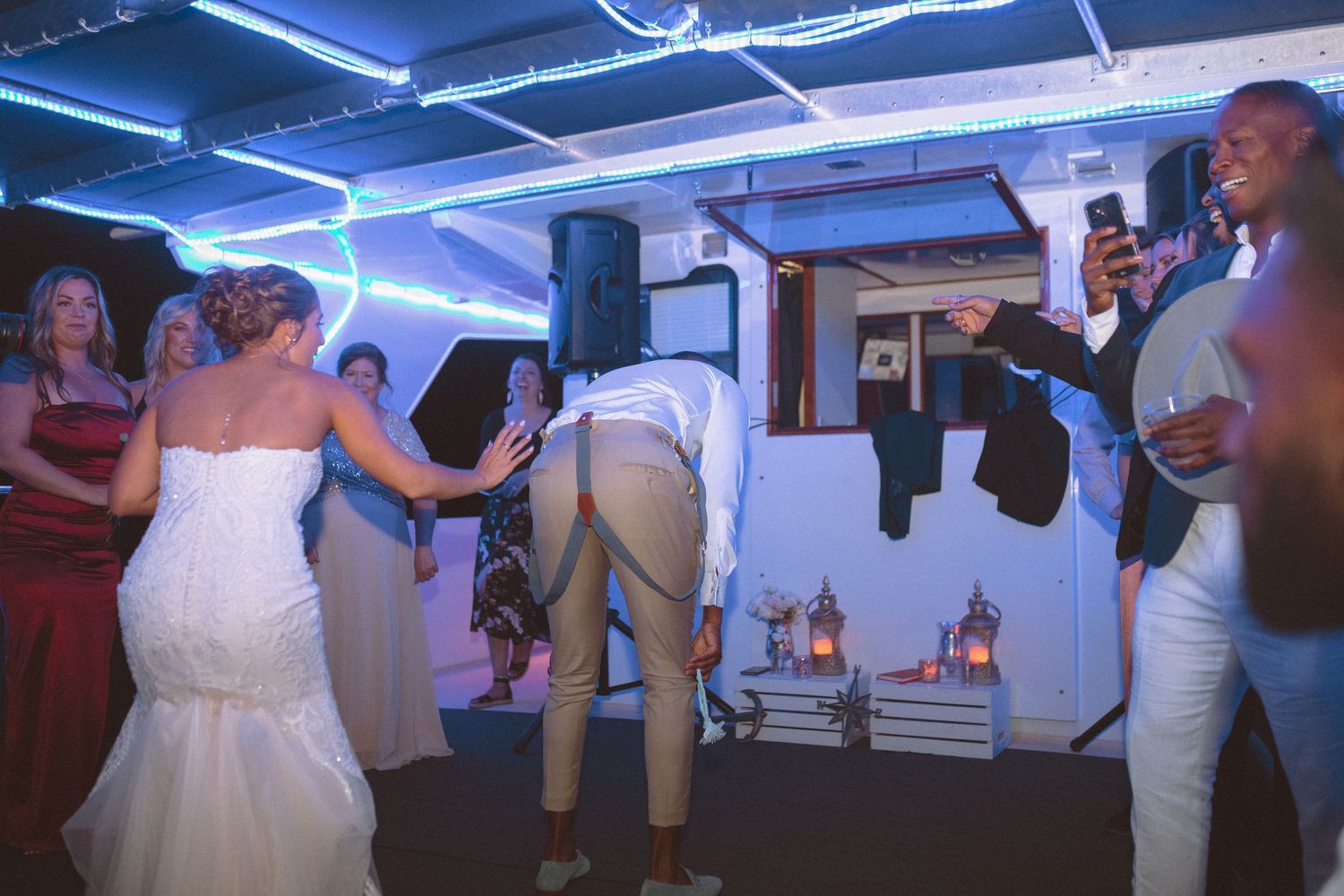 Groom bending over playfully while bride and guests laugh at wedding reception party.