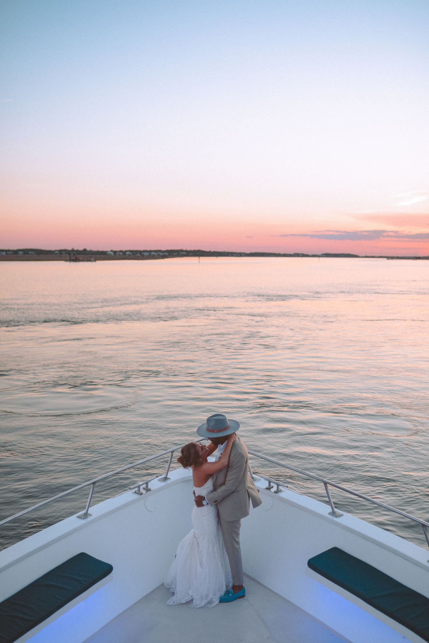 A bride and groom share a romantic kiss on the bow of a yacht at sunset over calm waters.