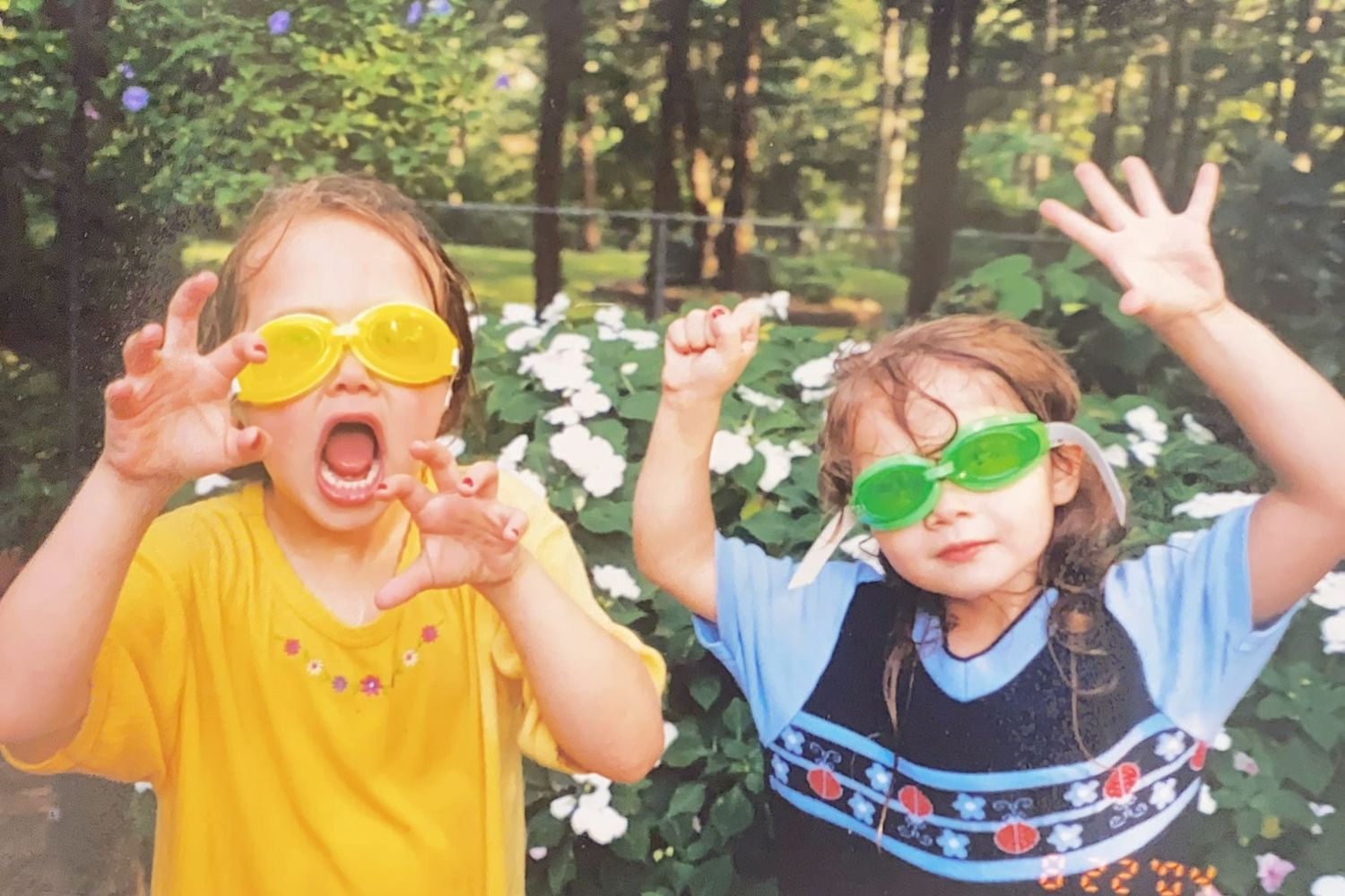 Two young girls wearing colorful oversized sunglasses make silly faces outdoors.