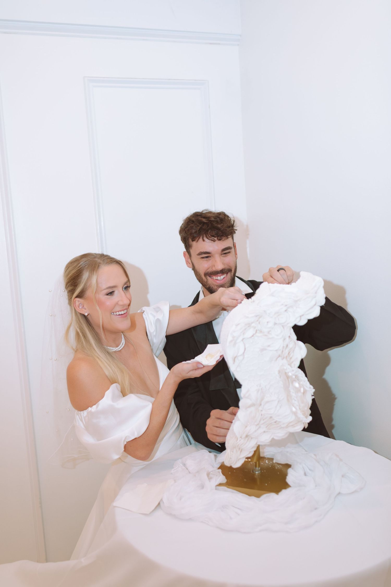 Smiling newlyweds cutting their ornate white floral wedding cake together at their reception.