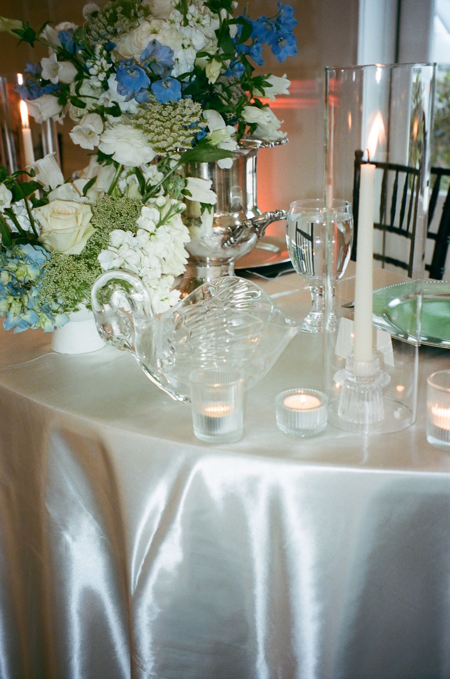 Elegant wedding table with white satin linen, crystal candle holders, tea lights, and blue and white floral centerpiece.
