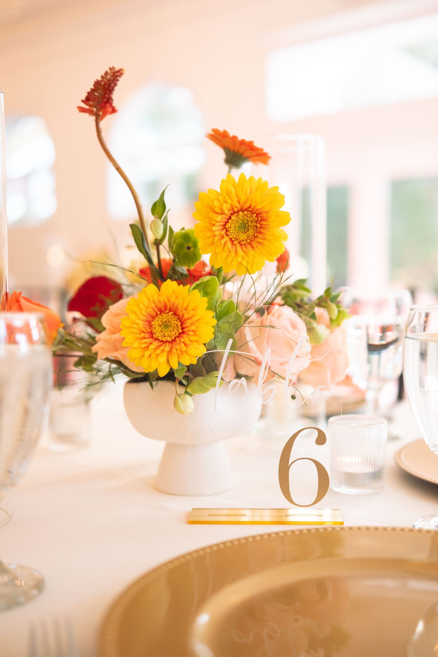 Vibrant yellow and orange gerbera daisy wedding centerpiece in white pedestal bowl with table number six.